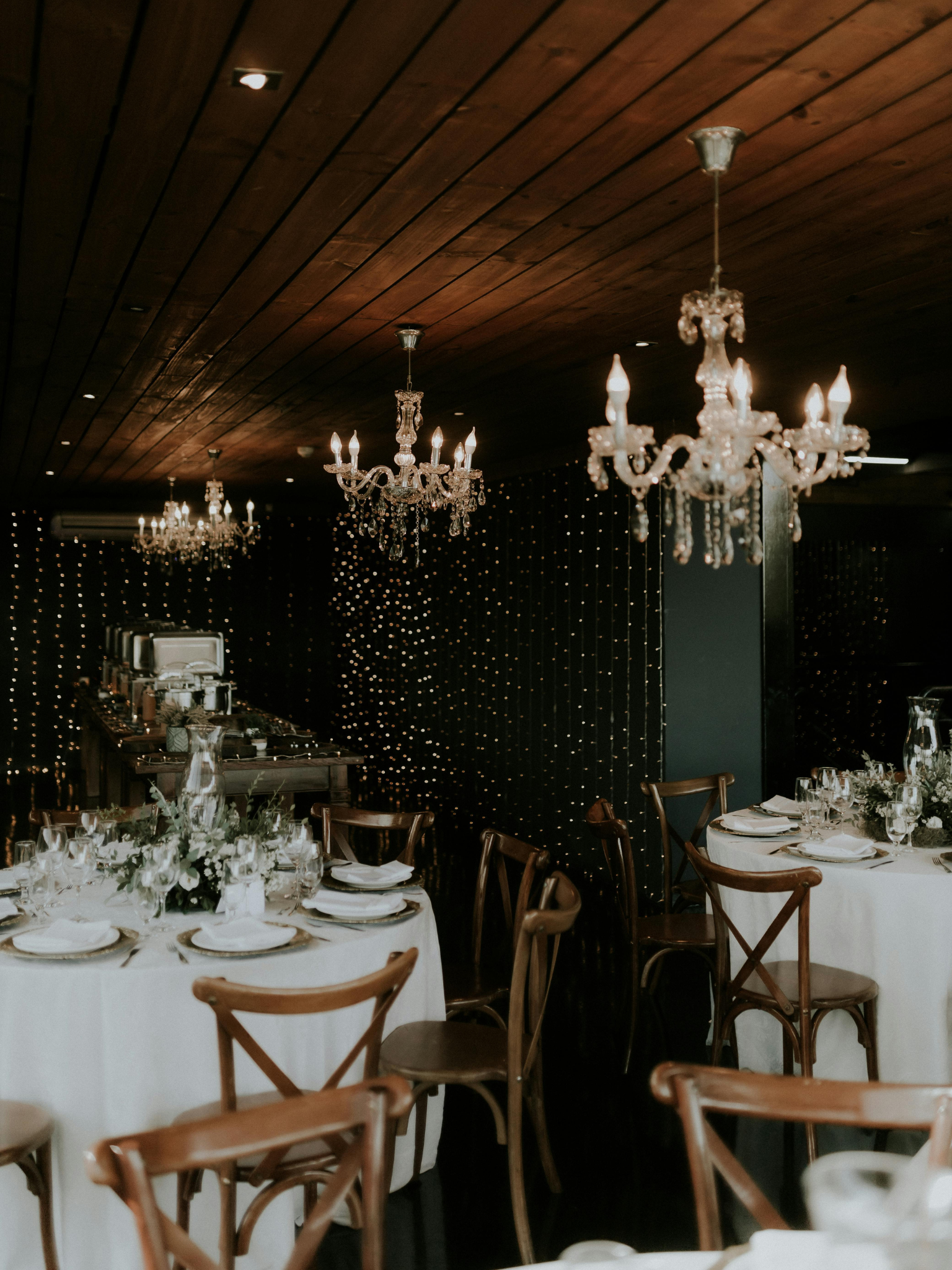 Decoración elegante en un salón con candelabros de cristal en el techo y mesas con manteles blancos, platos, cubiertos y arreglos florales para una celebración o evento.