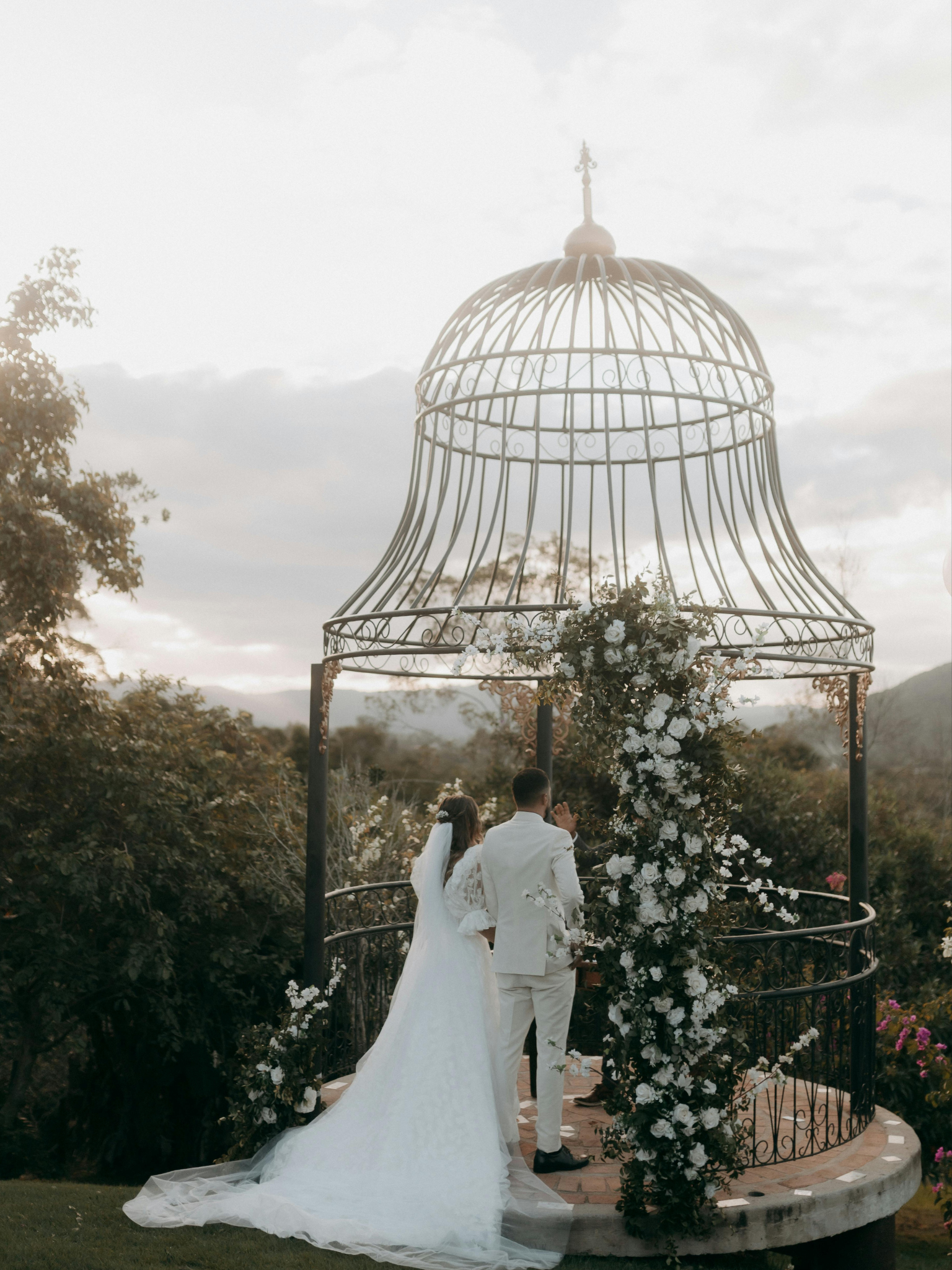 Una pareja de novios en una ceremonia de bodas en un entorno al aire libre con un quiosco de hierro decorado con flores blancas y verdes, al atardecer con un cielo nublado y montañas de fondo.
