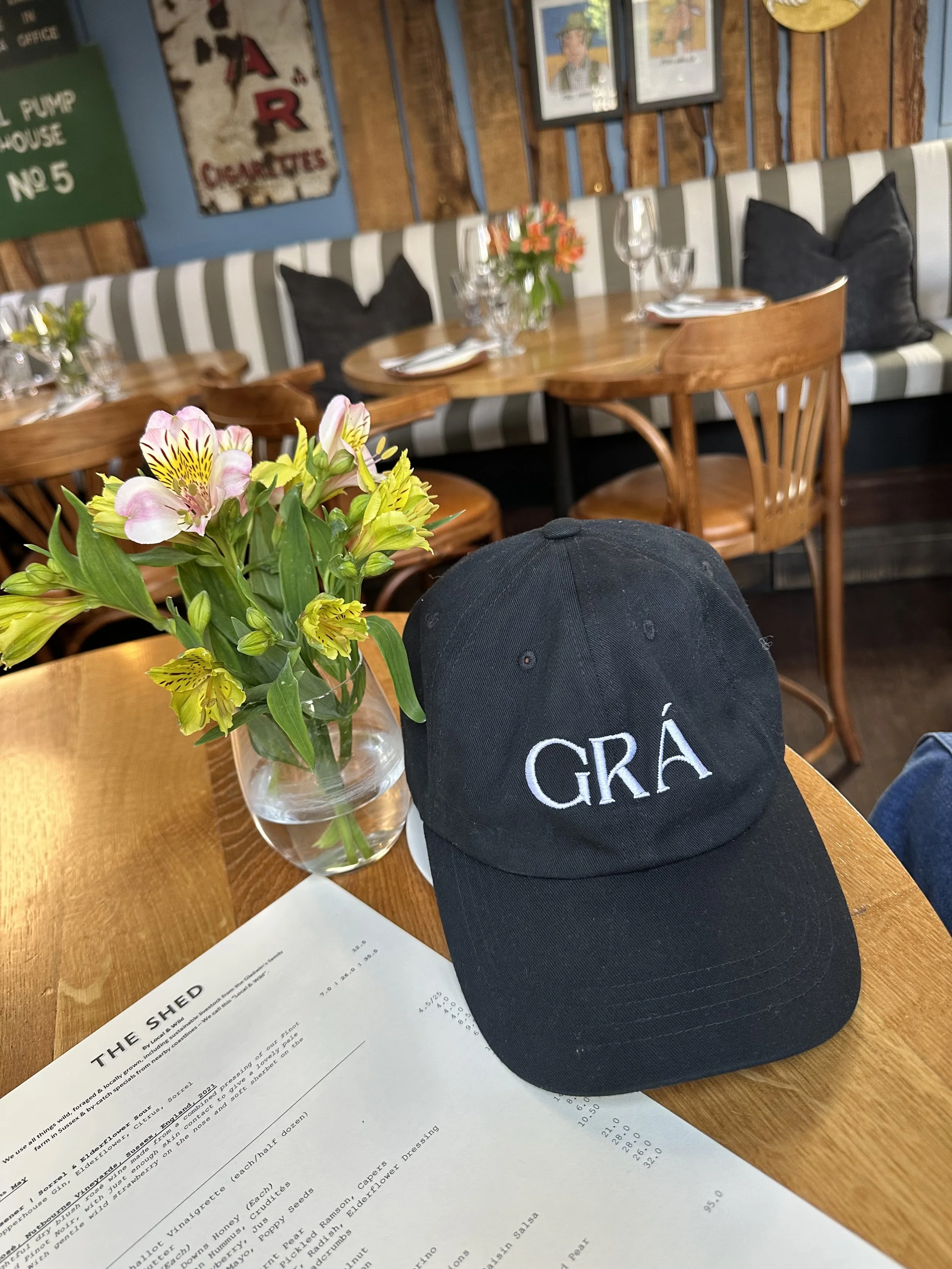 A black baseball cap with the word 'GRA' embroidered in white rests on a wooden table at SHED restaurant in Nottingham Hill, London. There is a vase with flowers, wine glasses, and silverware on the table. The environment is warm and eclectic.