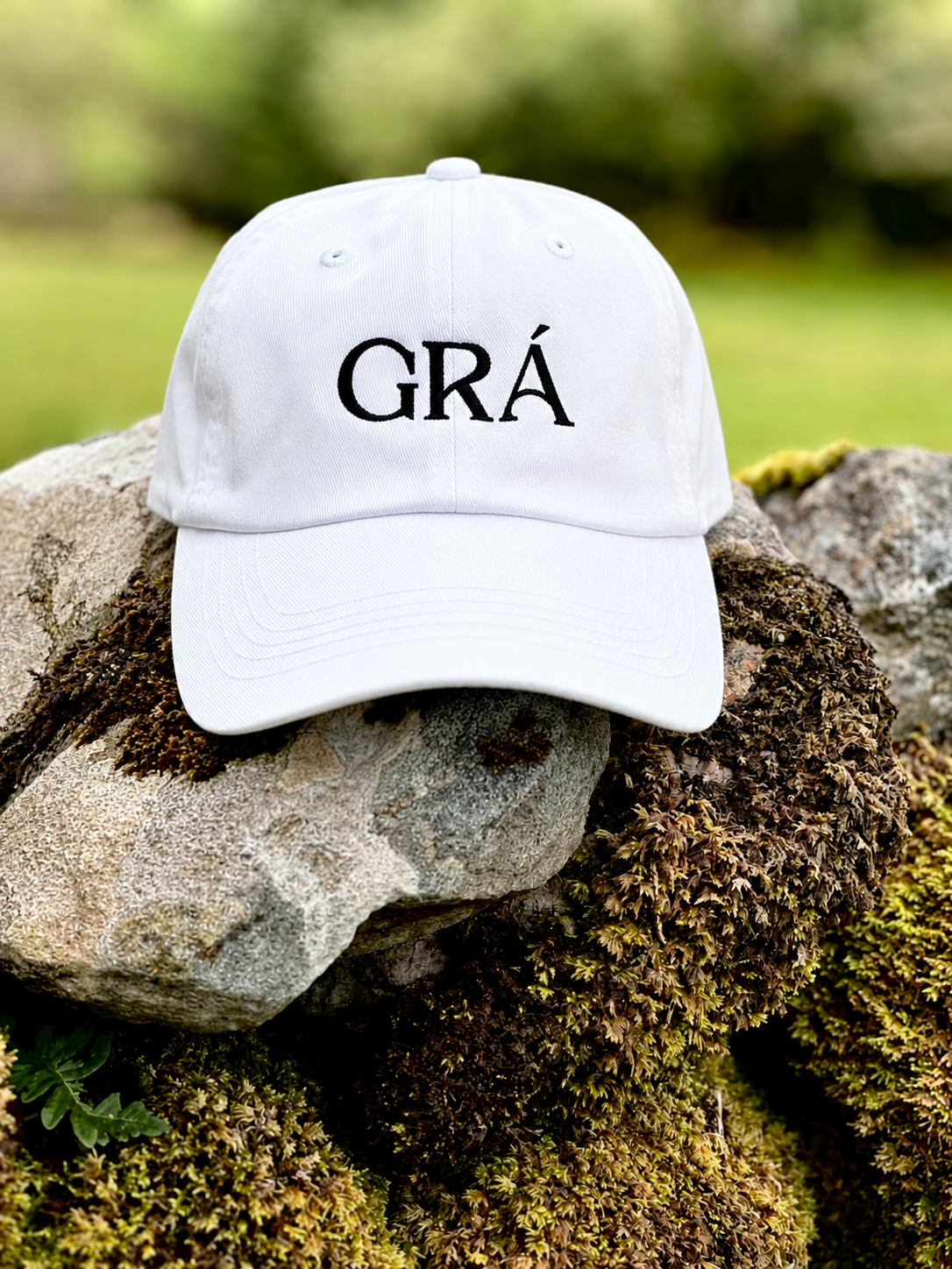 A white baseball cap with the word "GRÁ" embroidered in black, placed on rocks covered with moss outdoors.