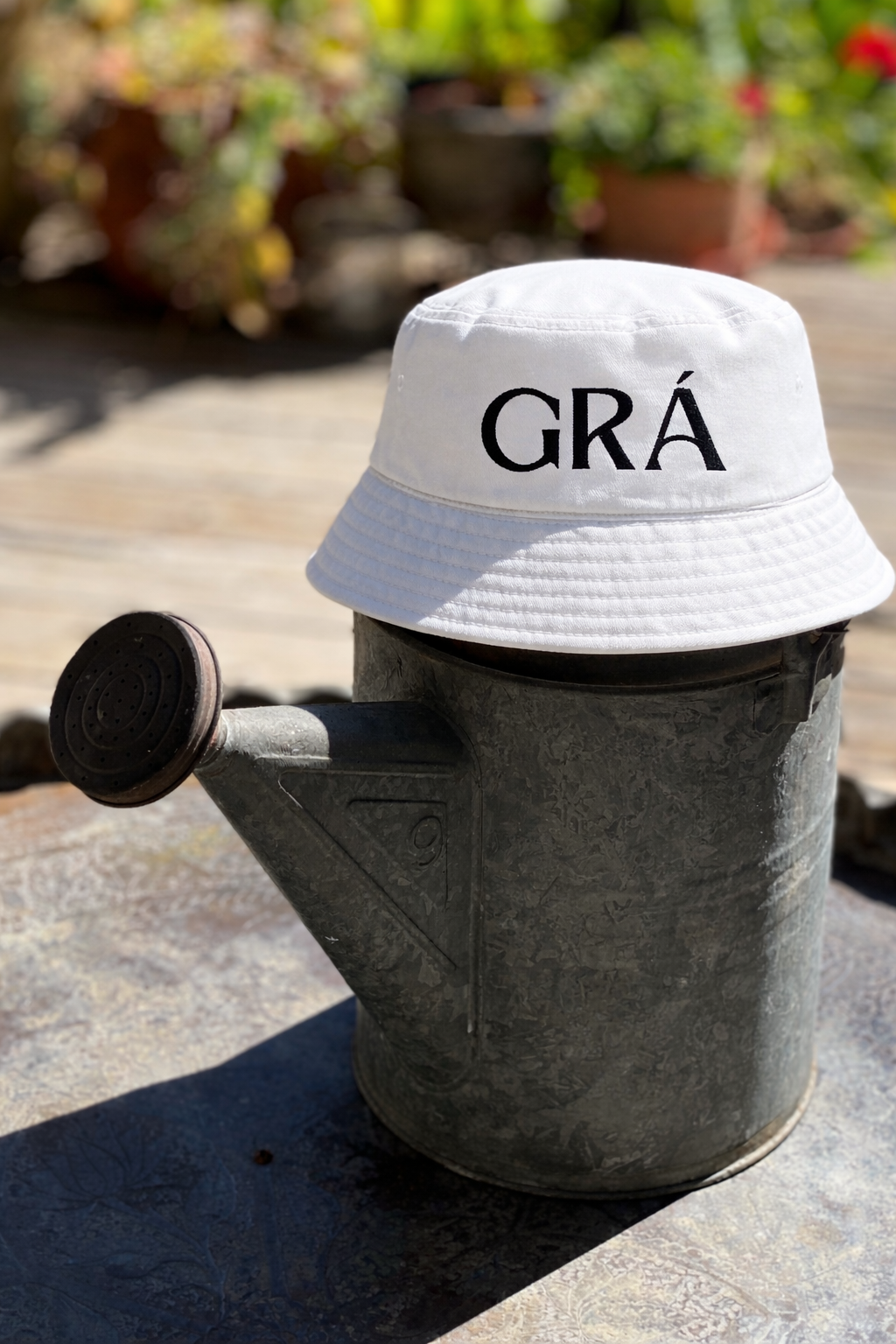 A white bucket hat with the word 'GRA' embroidered in black is placed on a gardening watering can. The watering can appears old and weathered, sitting outdoors on a stone surface with a blurred background of potted plants and greenery.