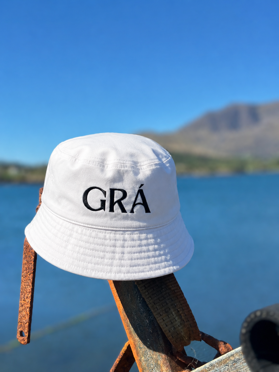 A white bucket hat with the word 'GRÁ' embroidered on it, resting on rusted metal, with a body of water and mountains in the background under a clear blue sky.