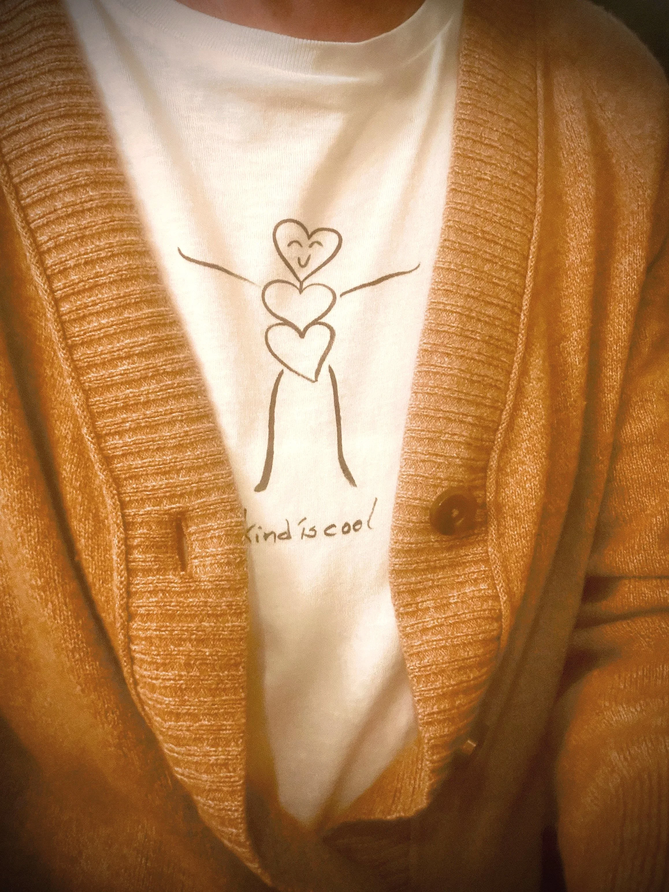 A person wearing a white t-shirt with a black line drawing of three hearts, smiling, and the words "kind is cool" beneath the drawing. The individual is also wearing a brown, ribbed cardigan.