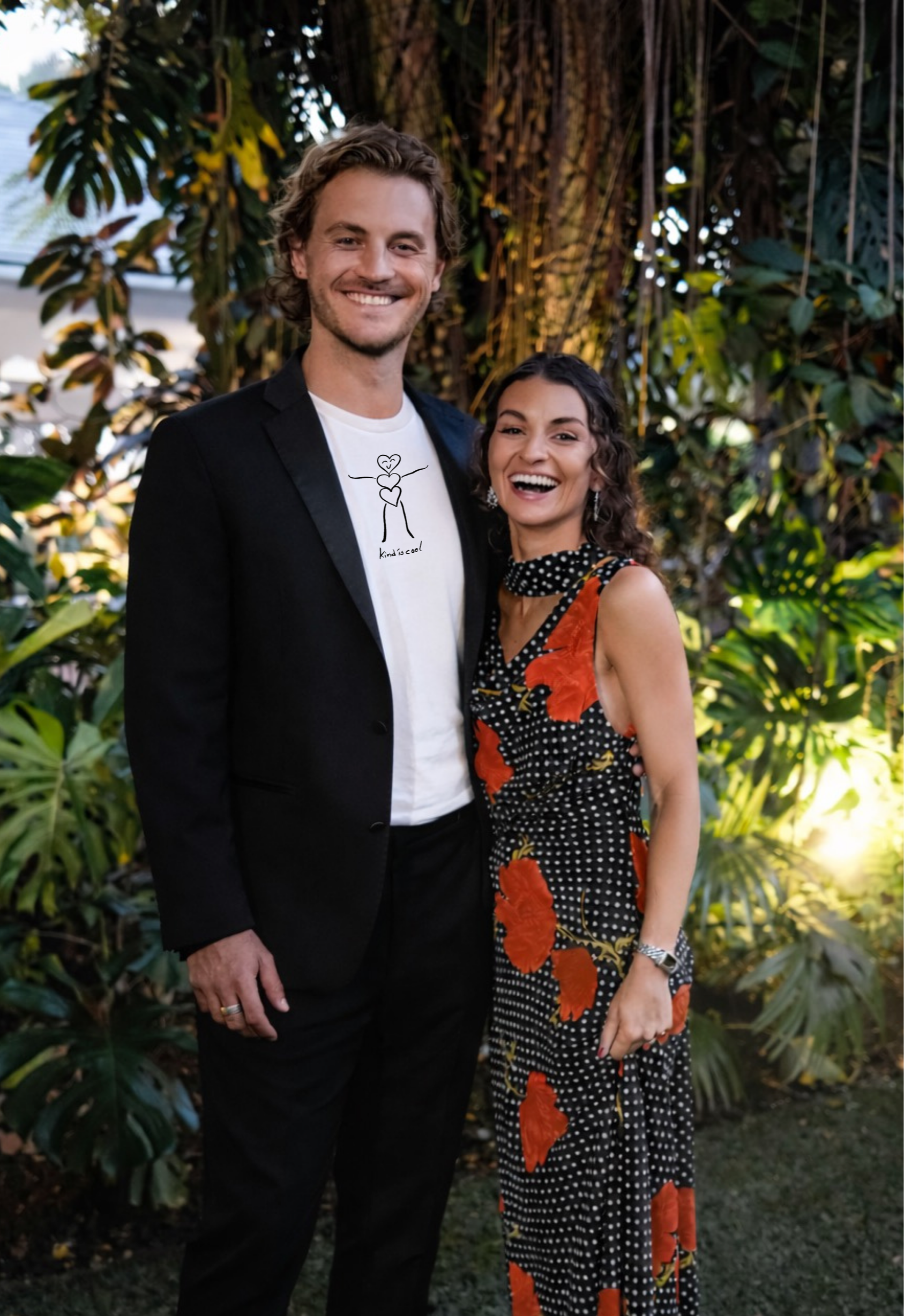 A man and a woman standing outdoors in front of lush green tropical plants, smiling at the camera. The man is wearing a black suit jacket over a white T-shirt with a minimalist line drawing and the word 'kindscool', and has wavy blonde hair. The woma
