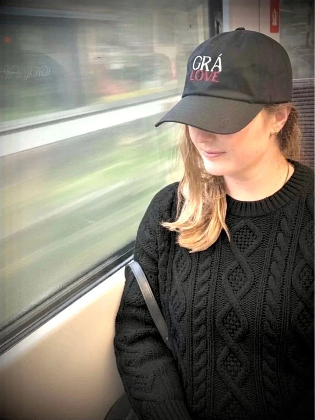 A woman wearing a black sweater and a black baseball cap with the words 'GRA LOVE' embroidered on it, sitting on a train with her eyes closed or looking down.