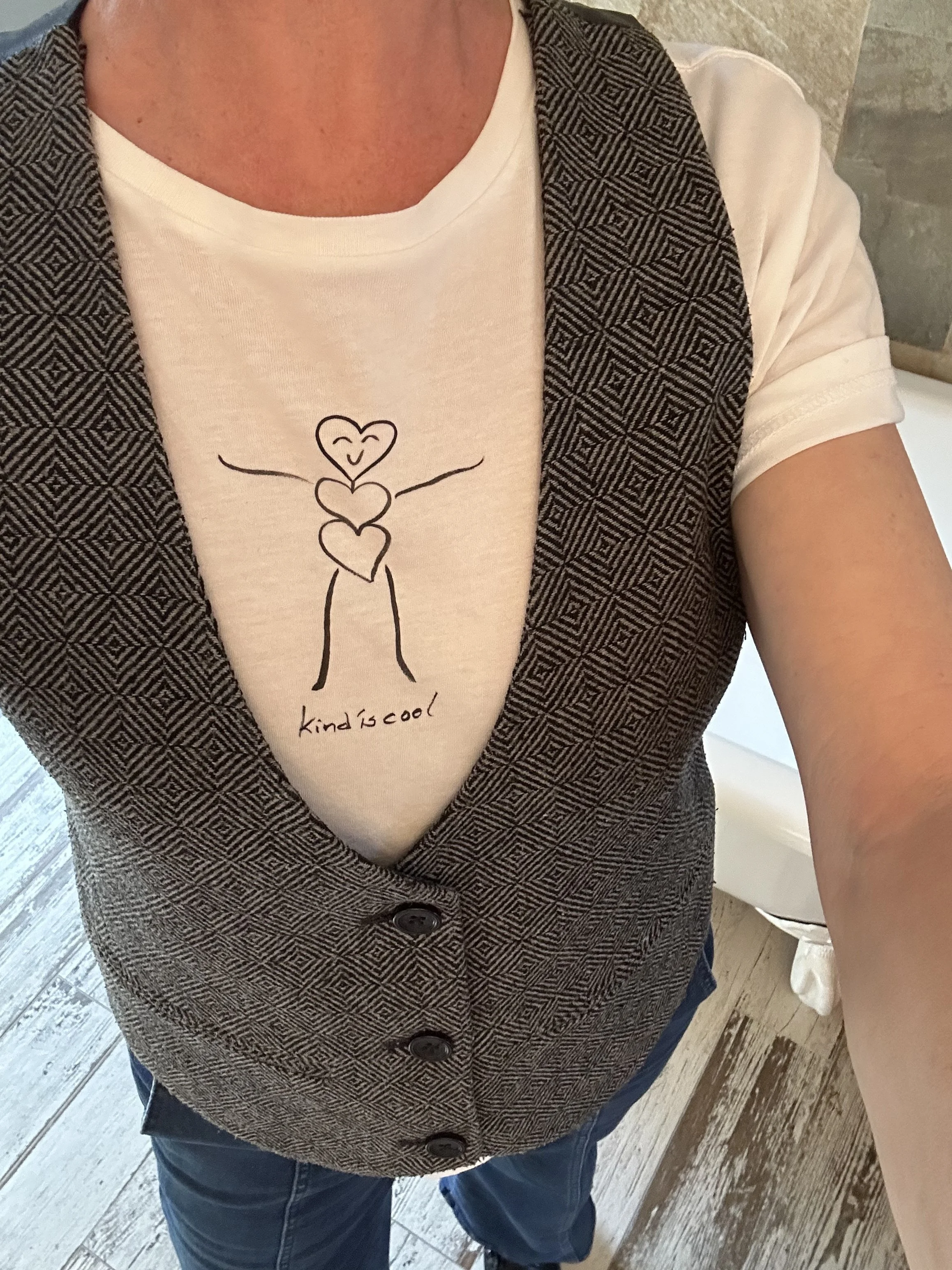 Person wearing a white t-shirt with a drawing of a smiling face and three hearts, a patterned vest, and blue jeans, taking a mirror selfie in a room with wooden flooring.