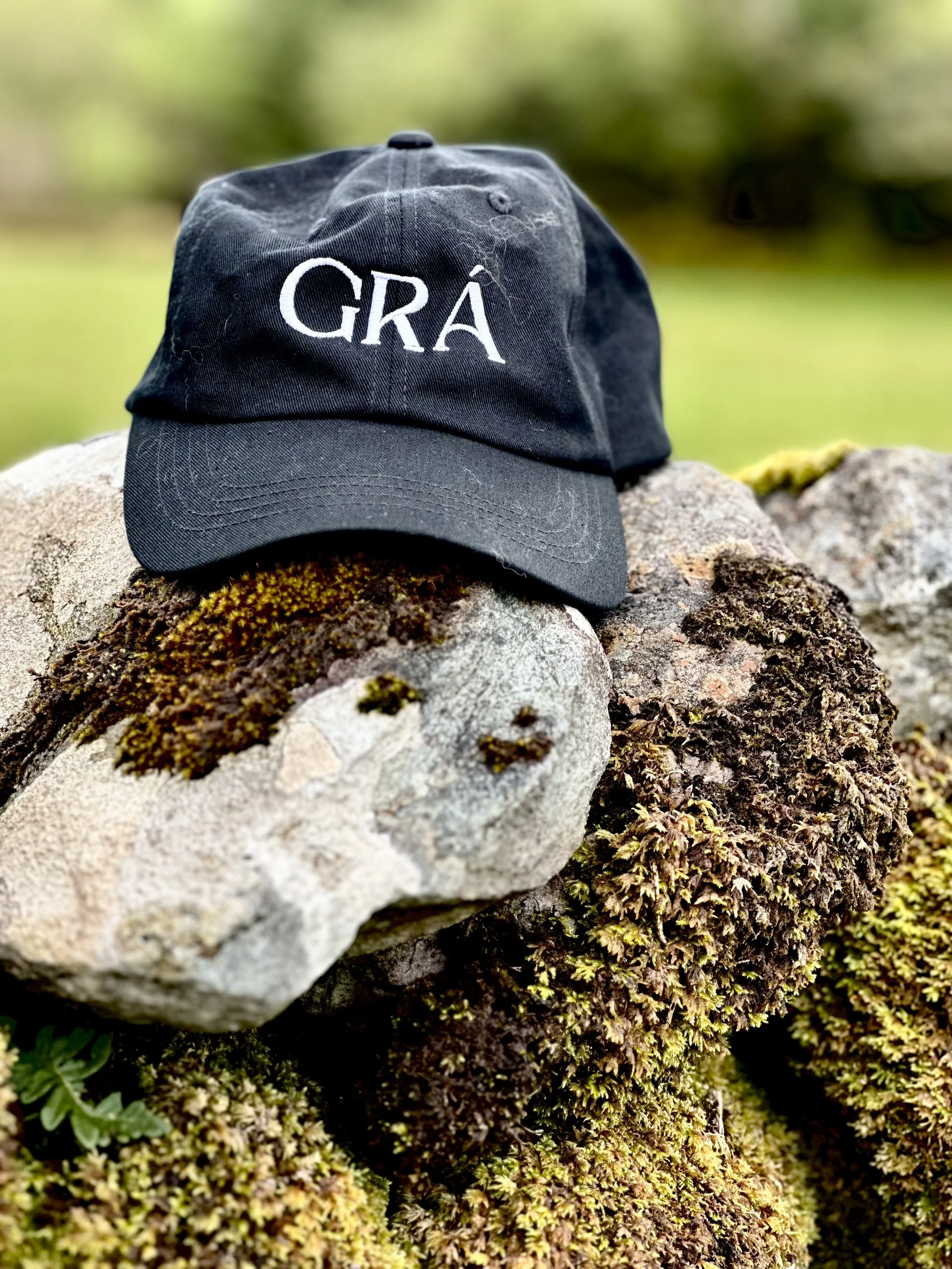 A black baseball cap with white embroidered text "GRA" resting on rocks and moss outdoors.