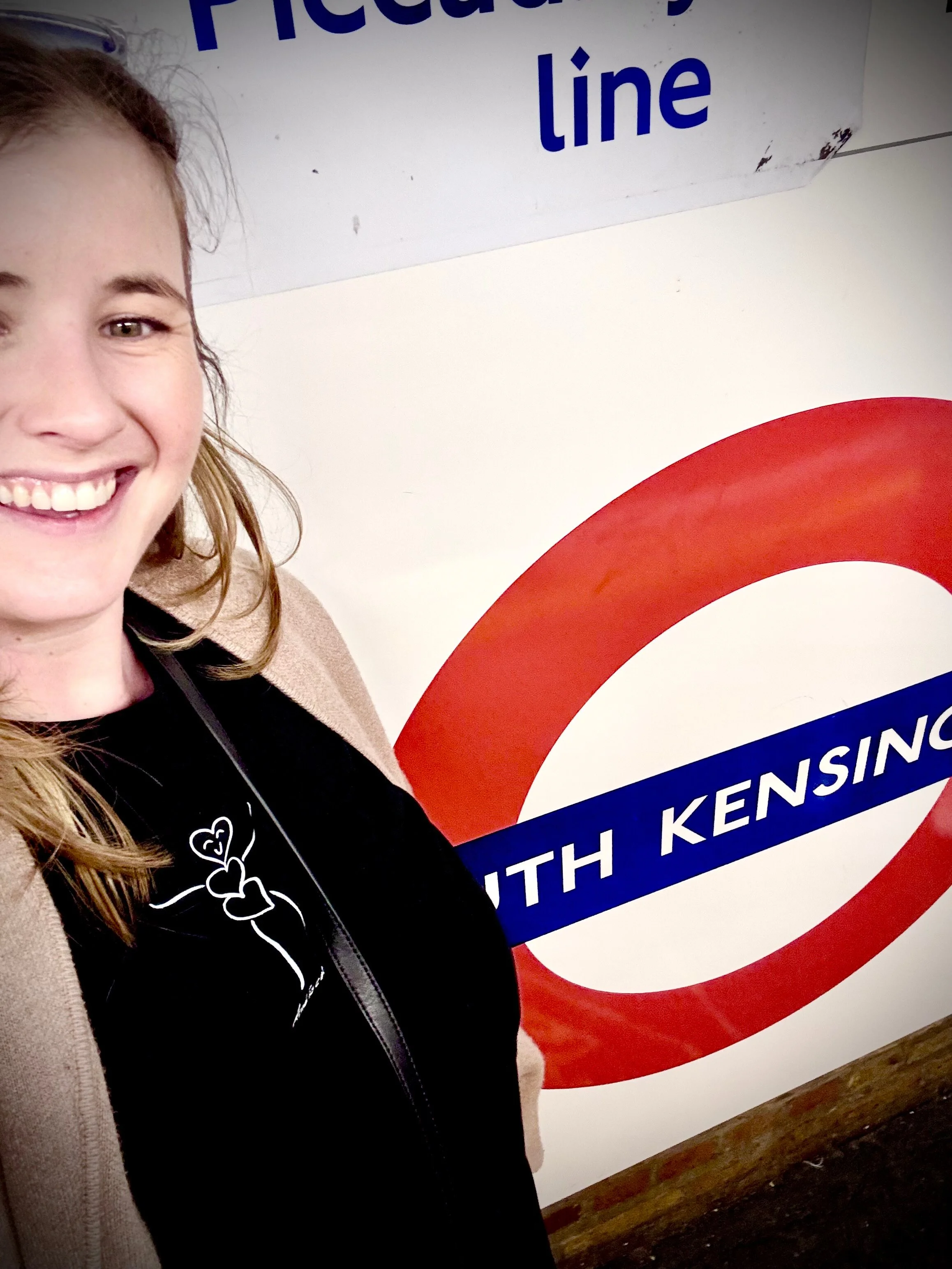 Half of a smiling woman's face and shoulder, standing in front of a London Underground sign with white background, red circle, blue bar with white text, partially visible.