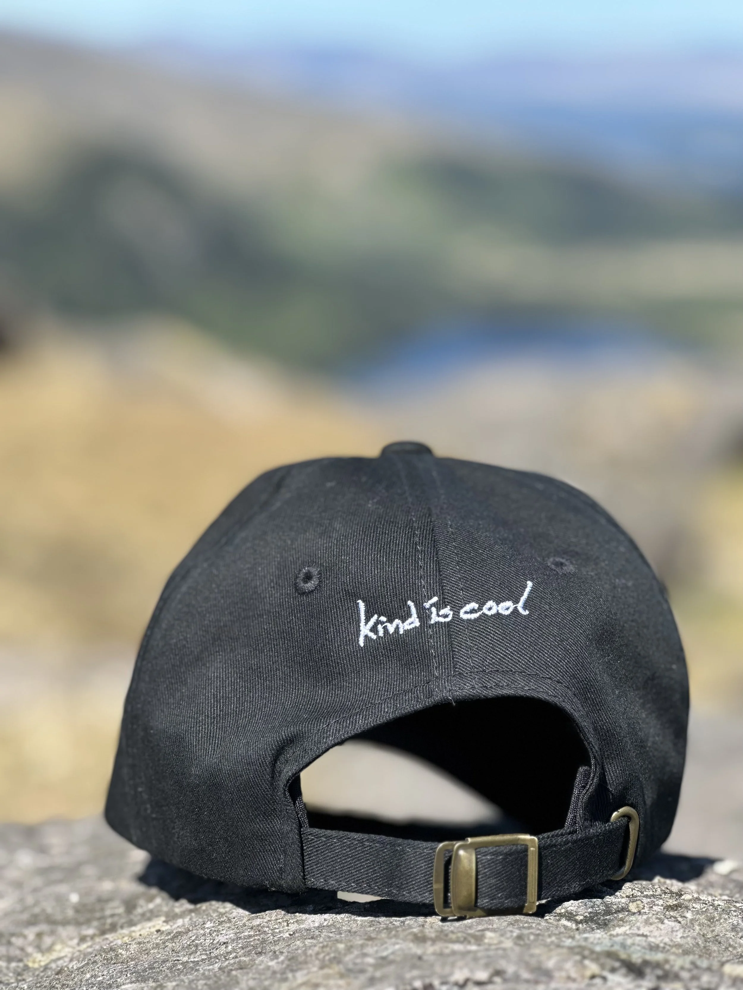 A black baseball cap with the phrase 'kind to cool' embroidered in white, placed on a rock outdoors with a blurred landscape in the background. The landscape is Ireland's Healy Pass between County Kerry and County Cork.