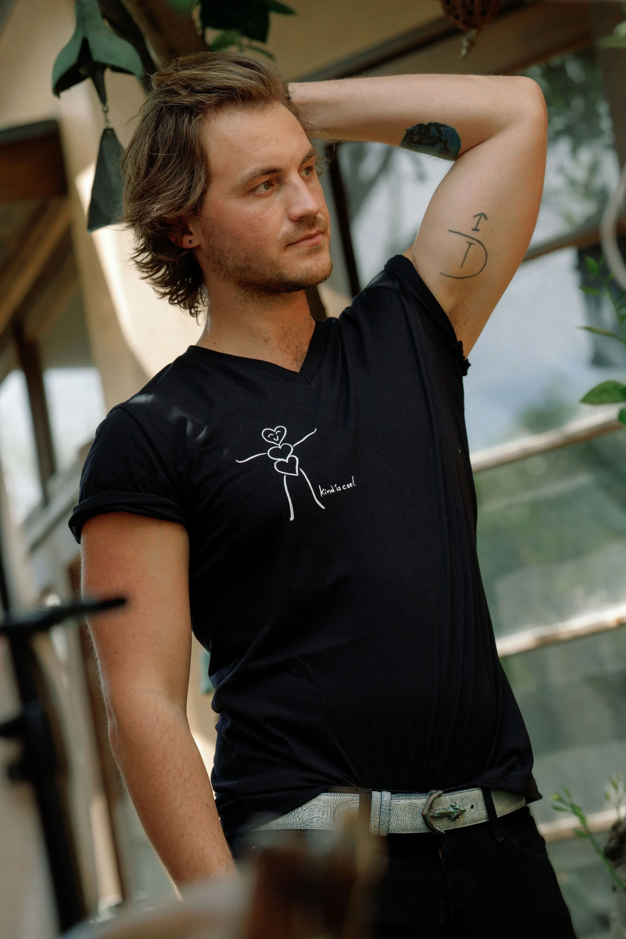 A young man with wavy brown hair posing with one arm raised behind his head. He has a tattoo on his inner arm and is wearing a black shirt with a small white graphic and text. The background is an indoor setting with plants and wooden shelves.