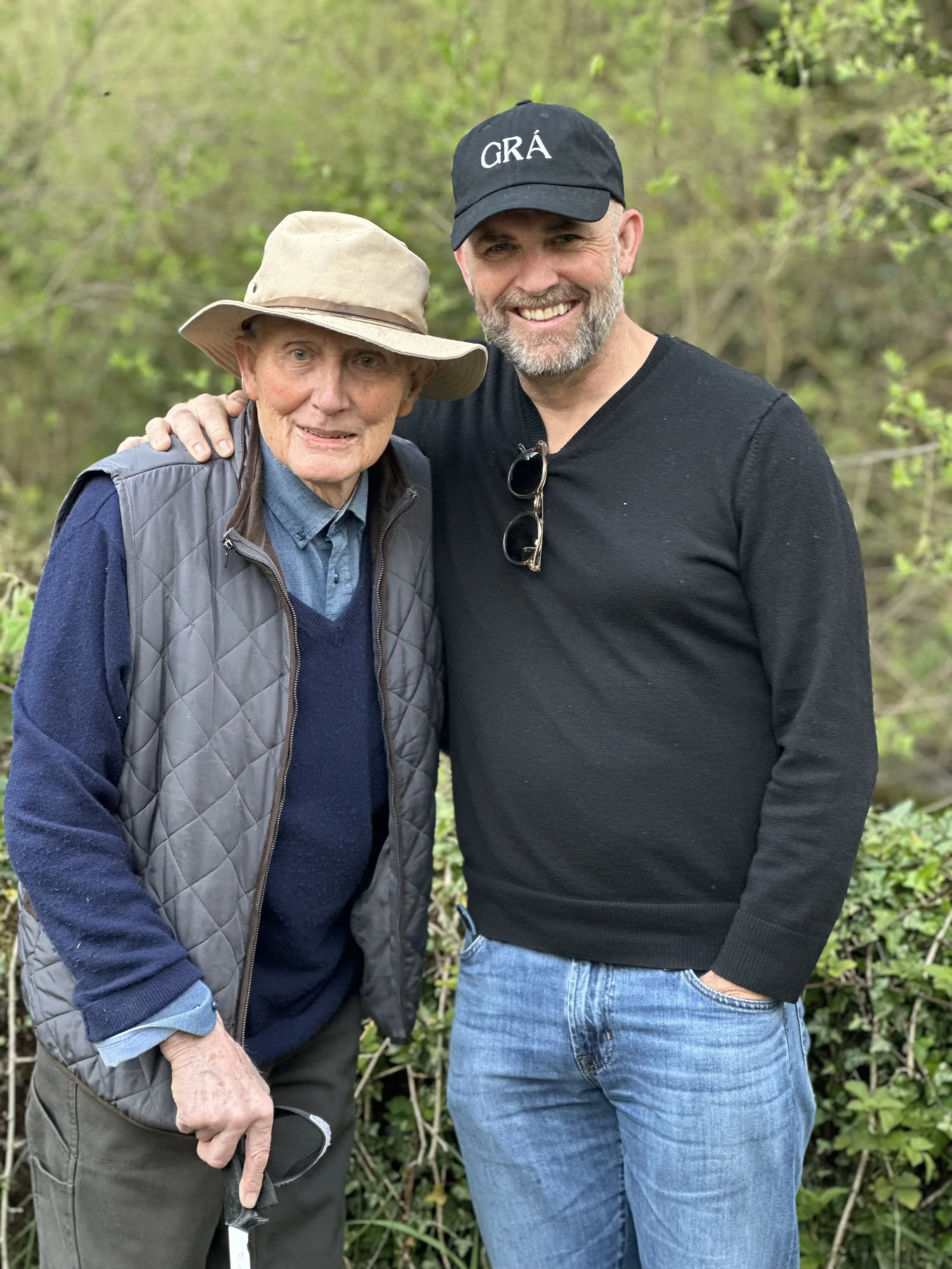 A smiling elderly man wearing a wide-brimmed hat and a navy blue vest next to a smiling man wearing a black cap with the word "GRA" , Love in Irish, on it and a black shirt, outdoors in a green, wooded area in Ballingeary, West Cork, Ireland.