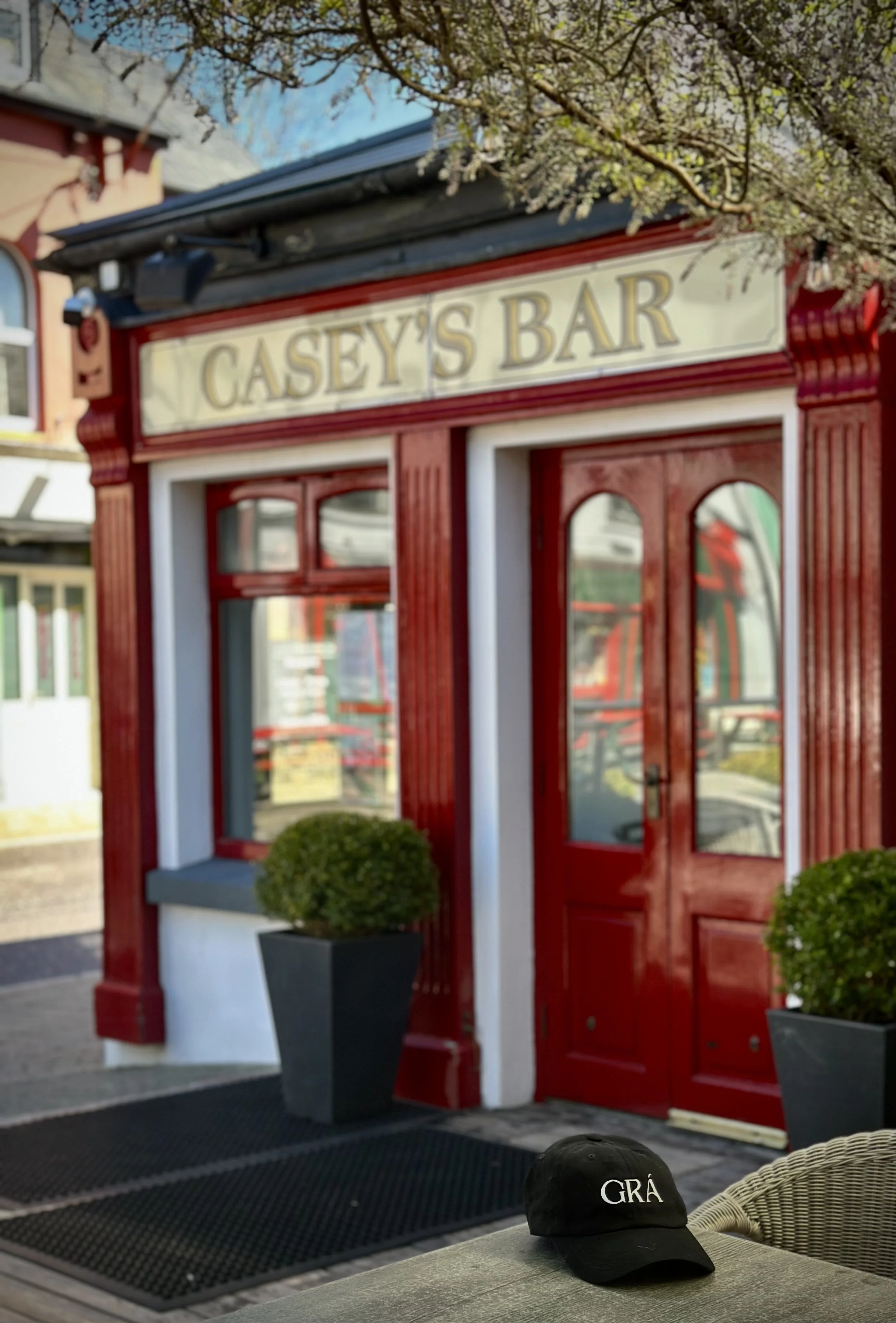 The entrance of Casey's Bar features a red exterior, large windows, and a sign above the door. There are various plants and vines. A black cap with the word 'GRA' is on a table in the foreground. Casey's is located in Glengarriff, West Cork, Ireland.