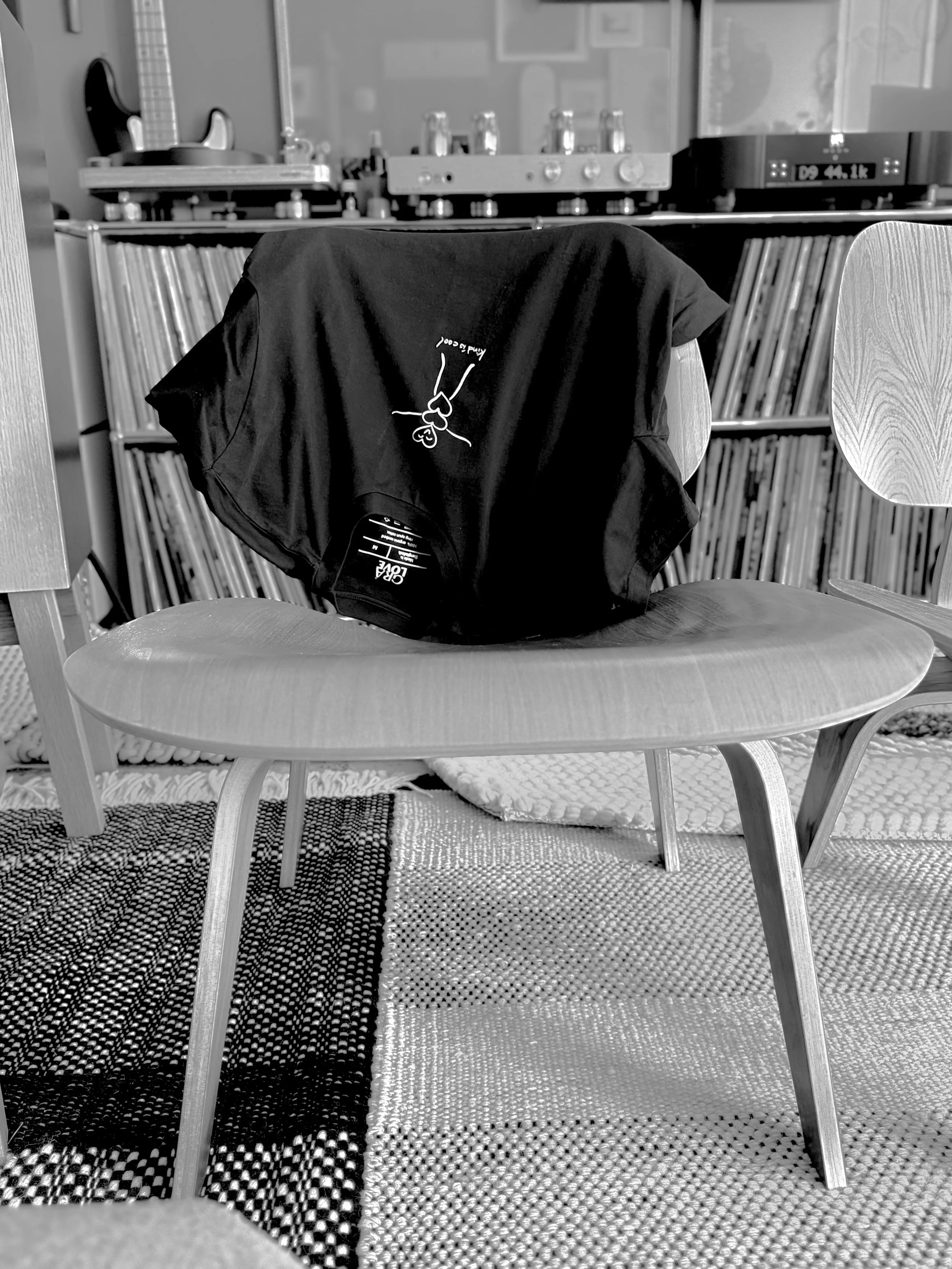 A black t-shirt with a white illustration of Grá Pal and the words, Kind is Cool, hangs over the back of a chair in front of a shelf filled with vinyl records, with a record player and a sound system on top of the shelf, located in Venice Beach, CA.
