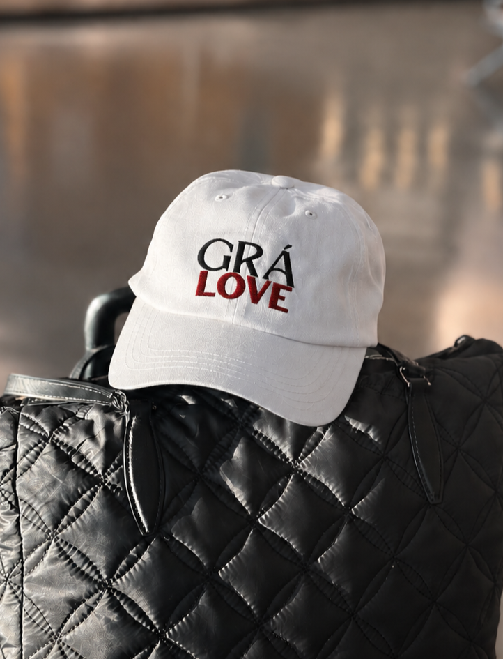 A white baseball cap with the words 'GRA LOVE' embroidered on the front in red and gray, placed on a wooden surface outdoors with a background of green foliage.