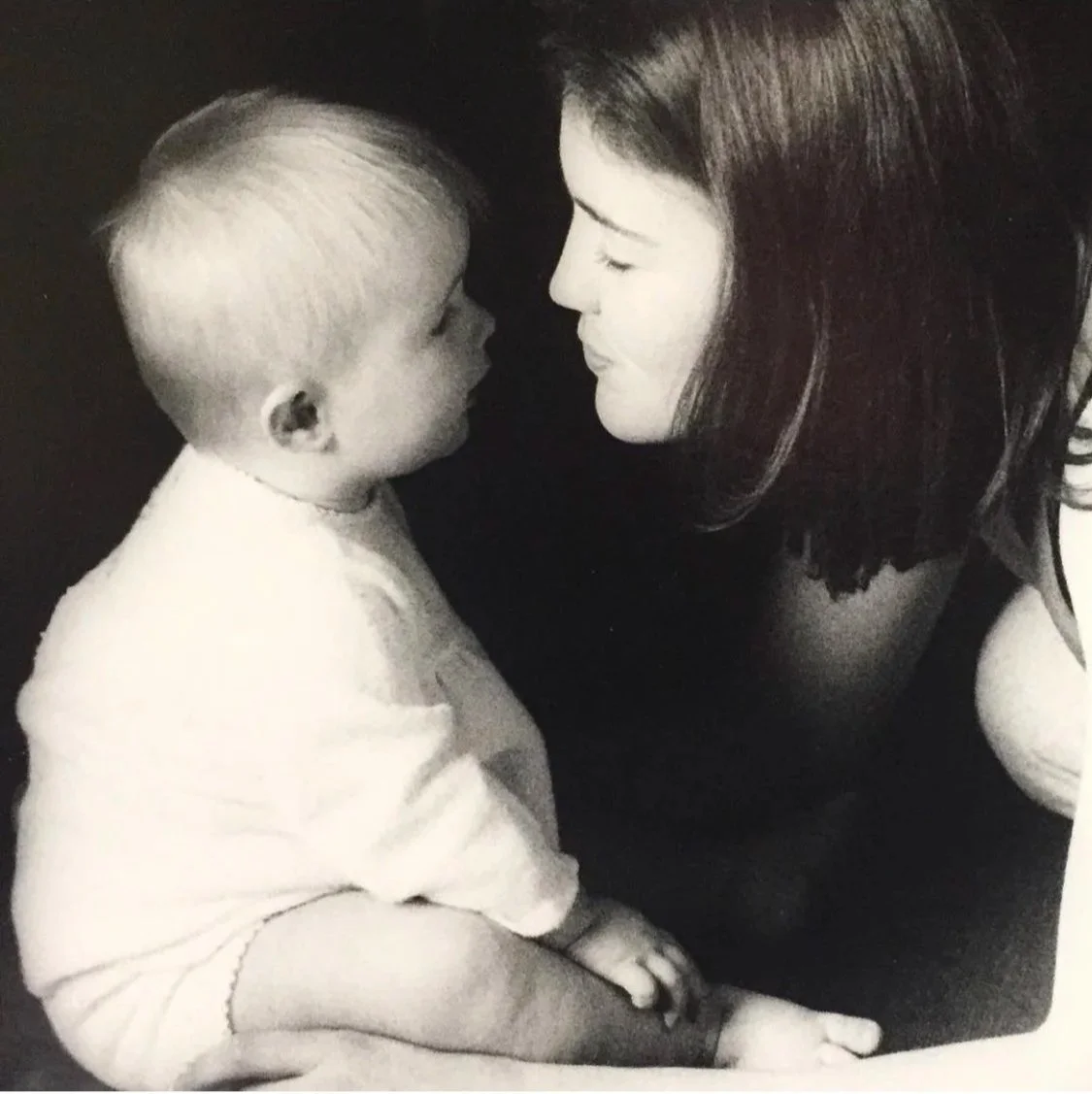 Black and white photo of a young woman and a toddler sitting facing each other with noses touching, eyes closed, sharing an intimate moment.