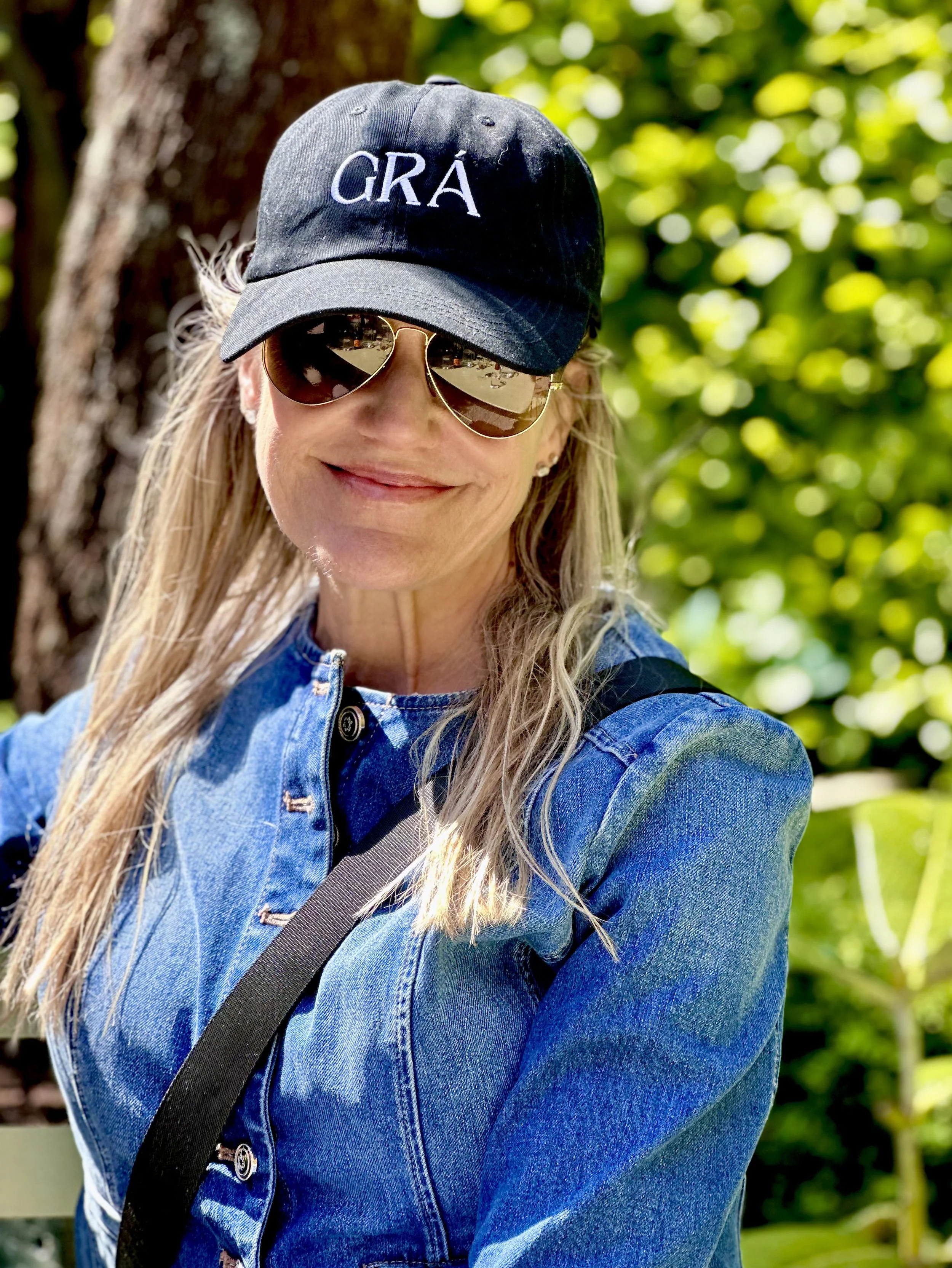 A woman with long blonde hair wearing a navy blue cap with white embroidered letters 'GRÁ'. She is also wearing sunglasses and a blue denim jacket, outdoors with blurred green foliage background at Dereen Garden Cafe, County Kerry, Ireland.