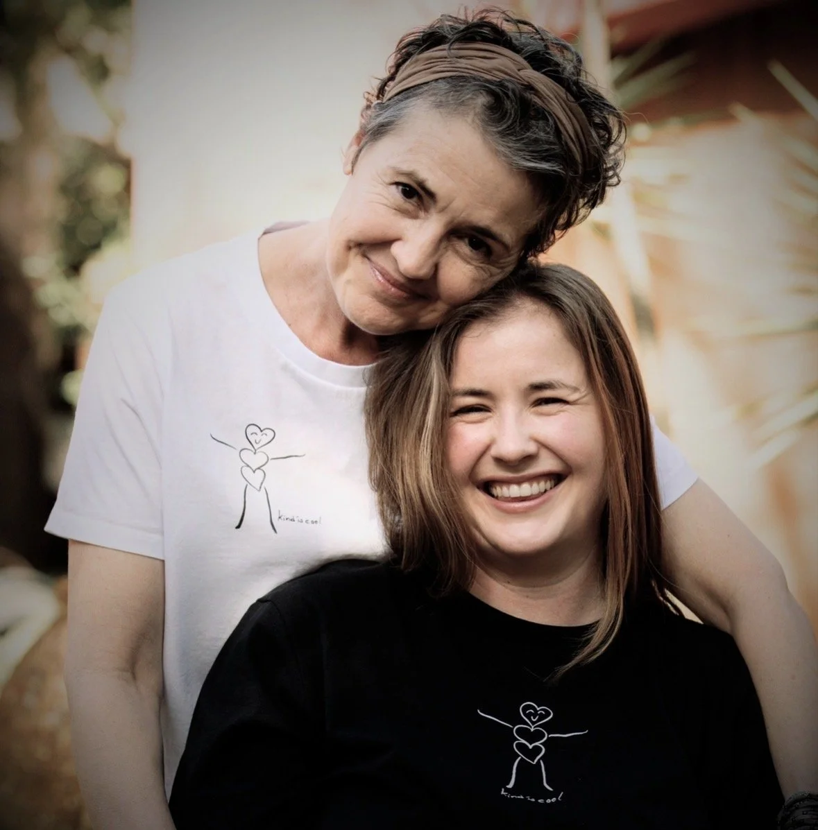 An older woman and a young woman smiling together outdoors, wearing matching t-shirts with a heart and stick figure drawing, with trees in the background.