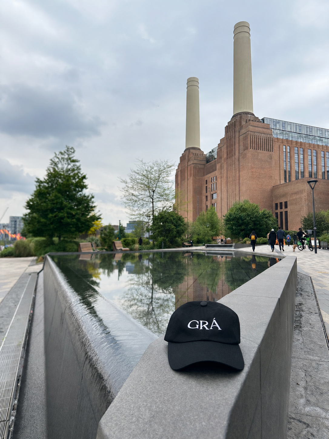 A black cap with the word 'GRÁ' placed on the edge of a modern water feature in front of Battersea Power Station, London. A large brick building with two tall smokestacks, surrounded by trees and people walking in an outdoor urban park.