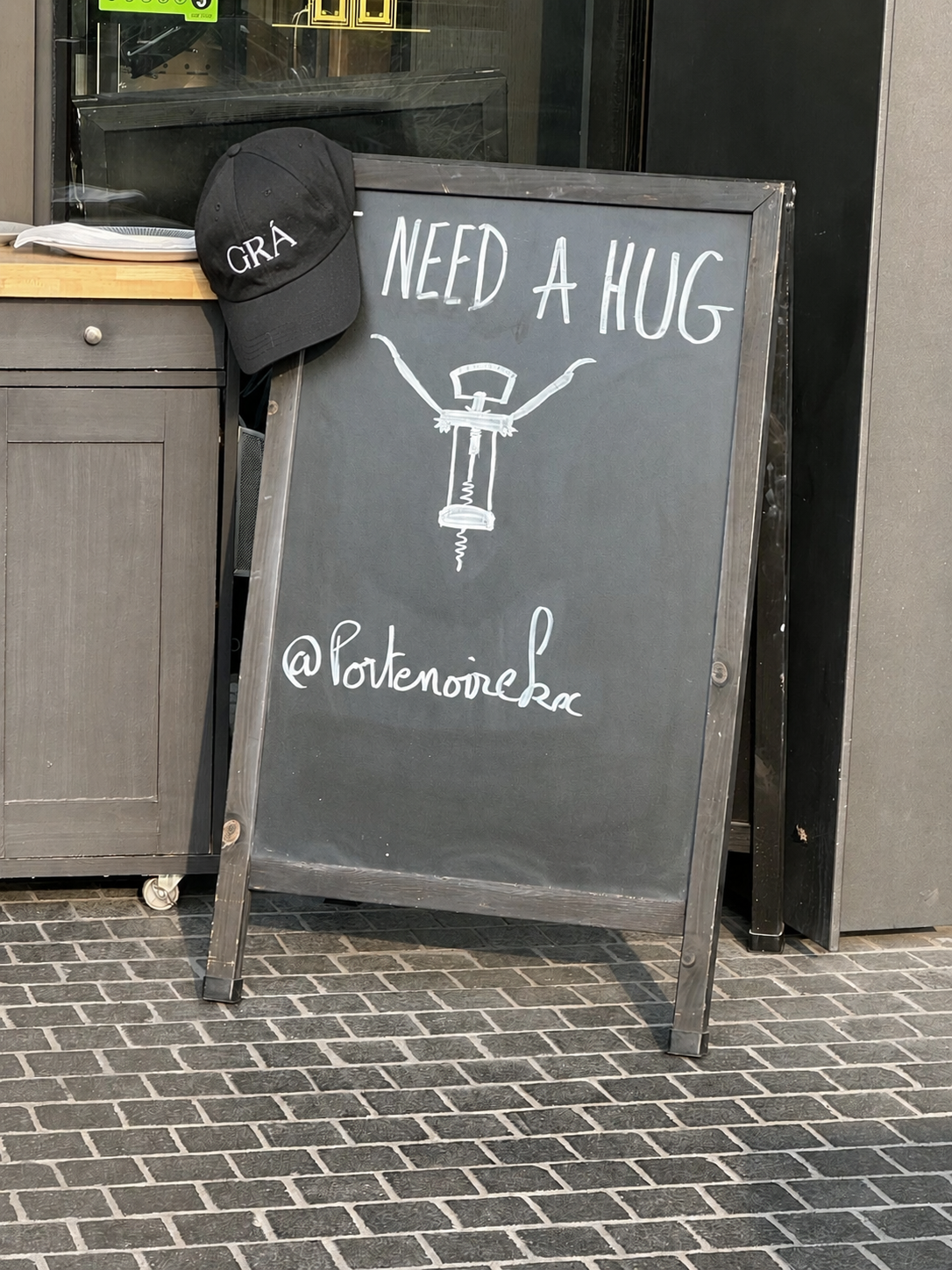 A chalkboard sign outside a store says 'Need a Hug' with a drawing of a screw and a handle, and includes the Instagram handle '@poutenoire.kc'. A black cap with 'GRA' embroidered on it rests on the corner of the sign in Coal Drop Yards, London, UK.