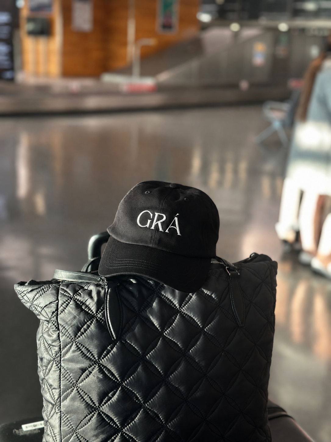 Black quilted jacket with a black cap featuring white embroidery reading "GRA" on it, sitting indoors at Cork Airport in County Cork, Ireland. 