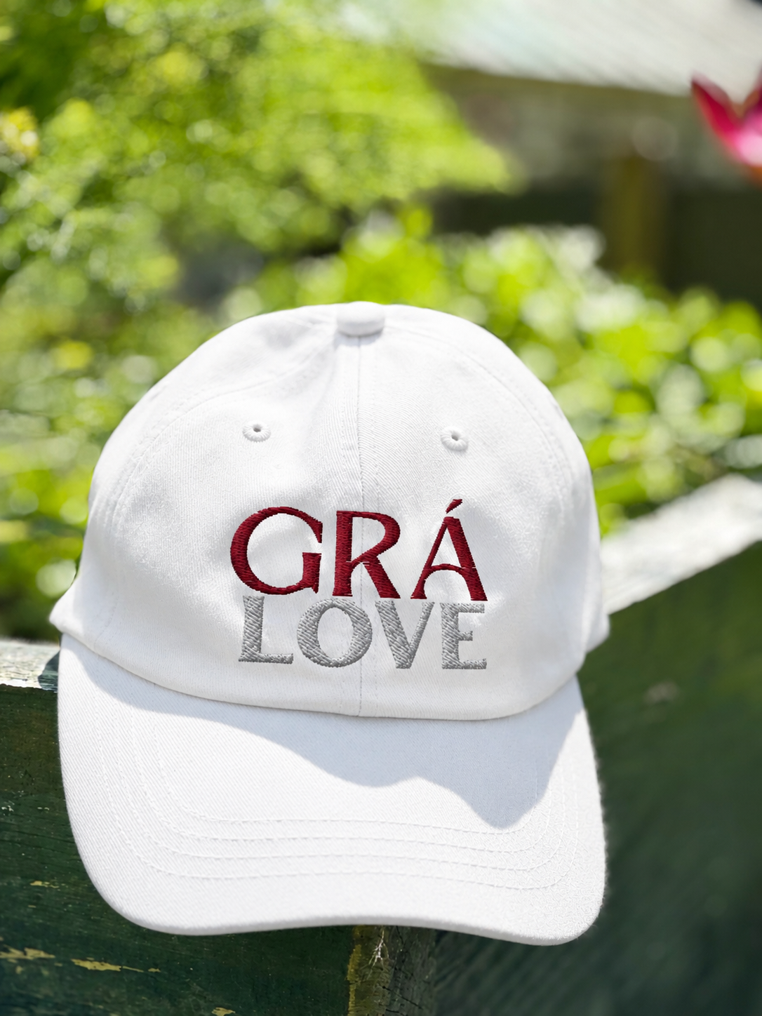 A white baseball cap with the words 'GRA LOVE' embroidered on the front in red and gray, placed on a wooden surface outdoors with a background of green foliage.