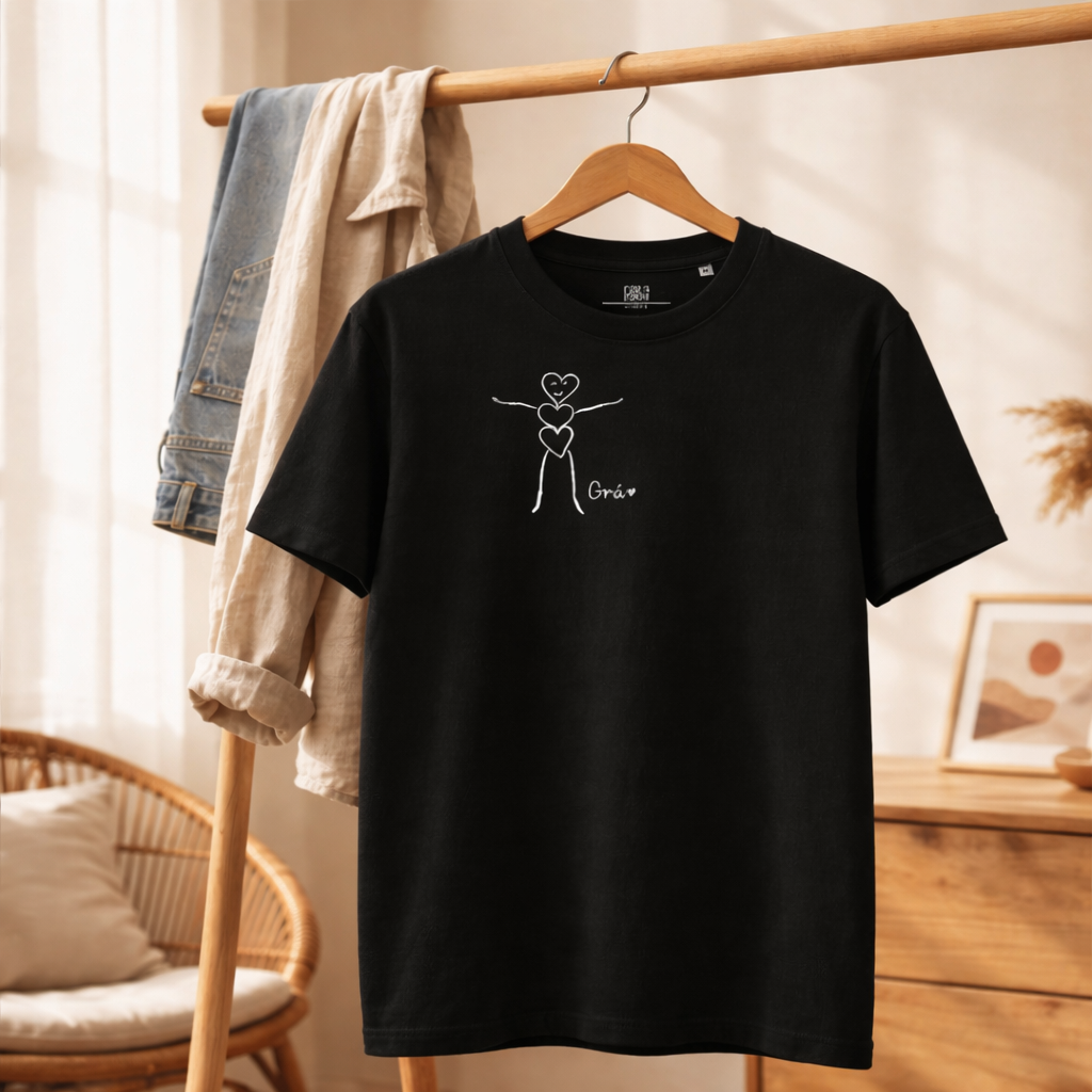 Black T-shirt with a white Japanese ink-style drawing of a smiling heart figure accompanied with the words, Kind is Cool. The shirt is hanging on a wooden hanger, with a beige and denim jacket hanging on a clothing rack behind it in a cozy room.