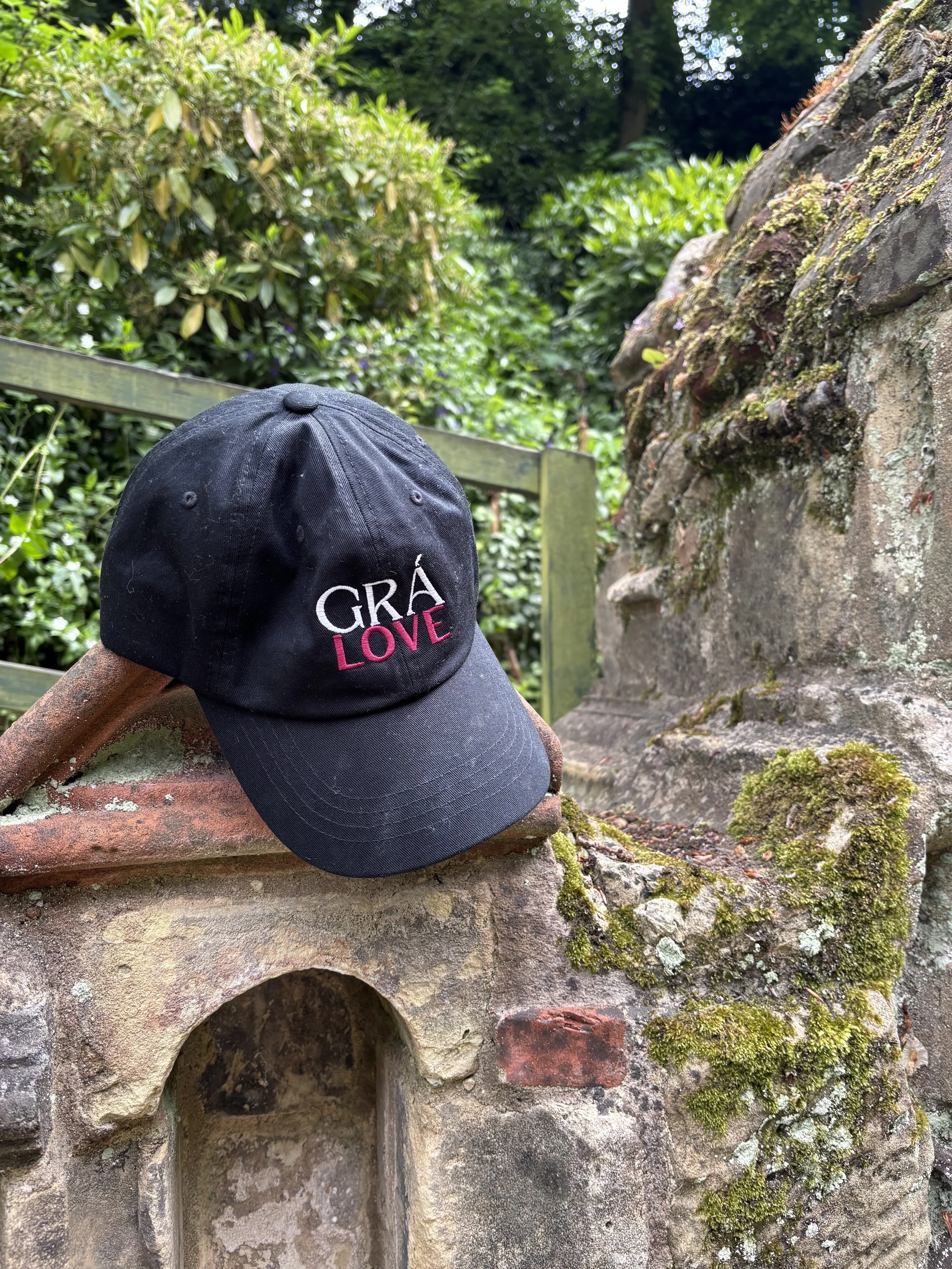 A black baseball cap with 'GRA LOVE' embroidered on the front, resting on a piece of brick and stone structure outdoors at the Plantation Garden in Norwich, London, England. Behind are green bushes and trees.