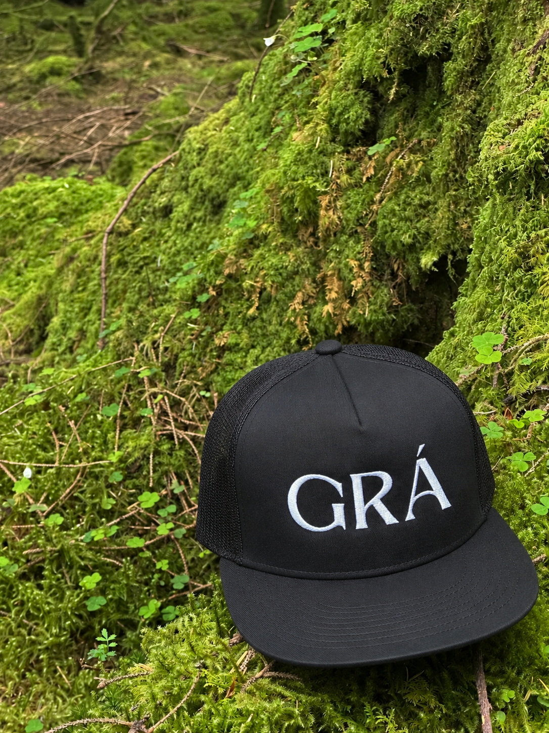 A black trucker hat with white embroidered text that reads 'GRA' resting on mossy ground in a forest setting.