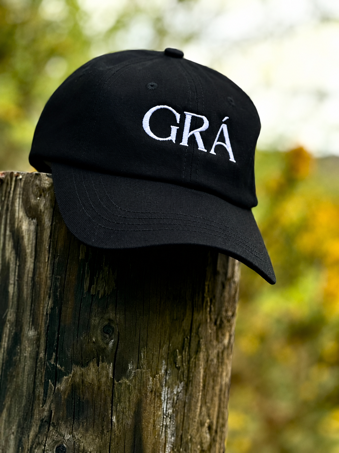 A black baseball cap with white embroidered text "GRA" resting on a weathered wooden post outdoors, with blurred green and yellow foliage in the background.