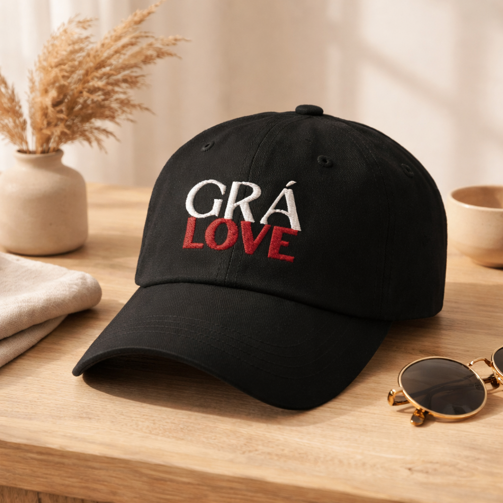 A classic black cotton cap with antique brass closure with the words "GRÁ LOVE" embroidered on the front in White and Deep Red. It sits next to a pair of round sunglasses, a cream-colored cloth, and a small beige vase with dried pampas grass.