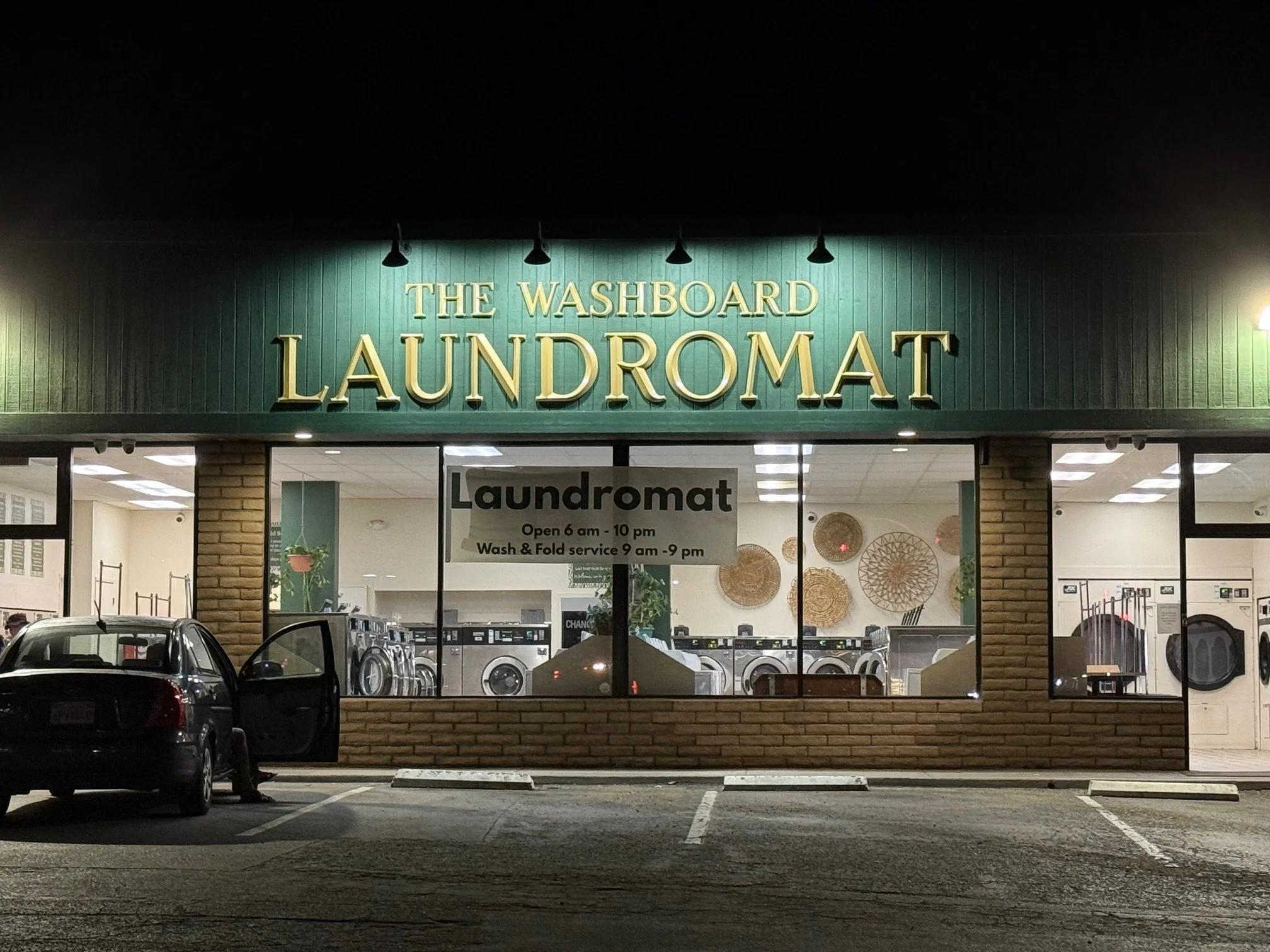 Night view of a laundromat with illuminated sign that reads 'The Washboard Laundromat', large front window showing washing machines inside, and a parked black car in front.