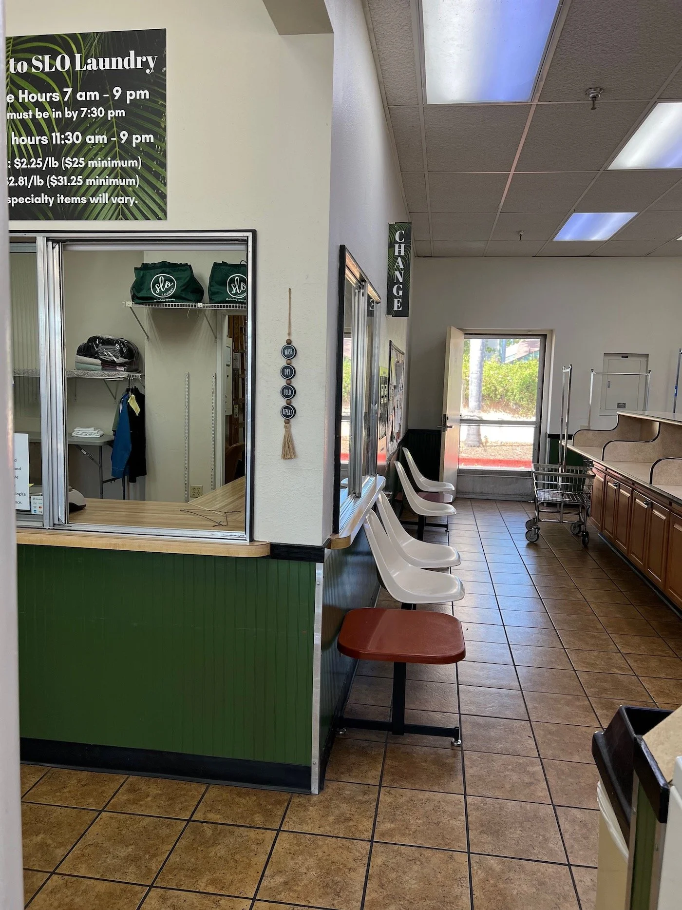 Inside a laundromat with white walls, a green countertop, row of white chairs, a shopping cart, and a window with a view outside. Sign with laundry hours on the wall.