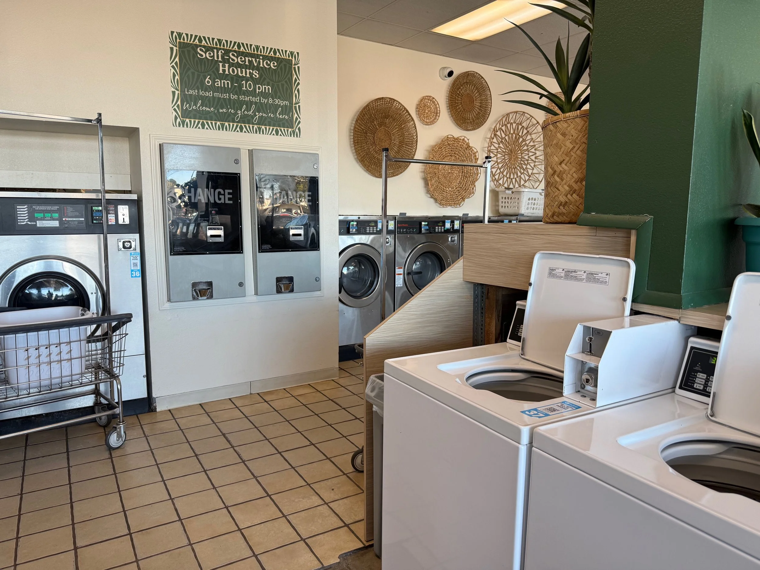 Laundry room with washers and dryers, a sign displaying self-service hours from 6 am to 10 pm, decorative wall baskets, and a green potted plant.