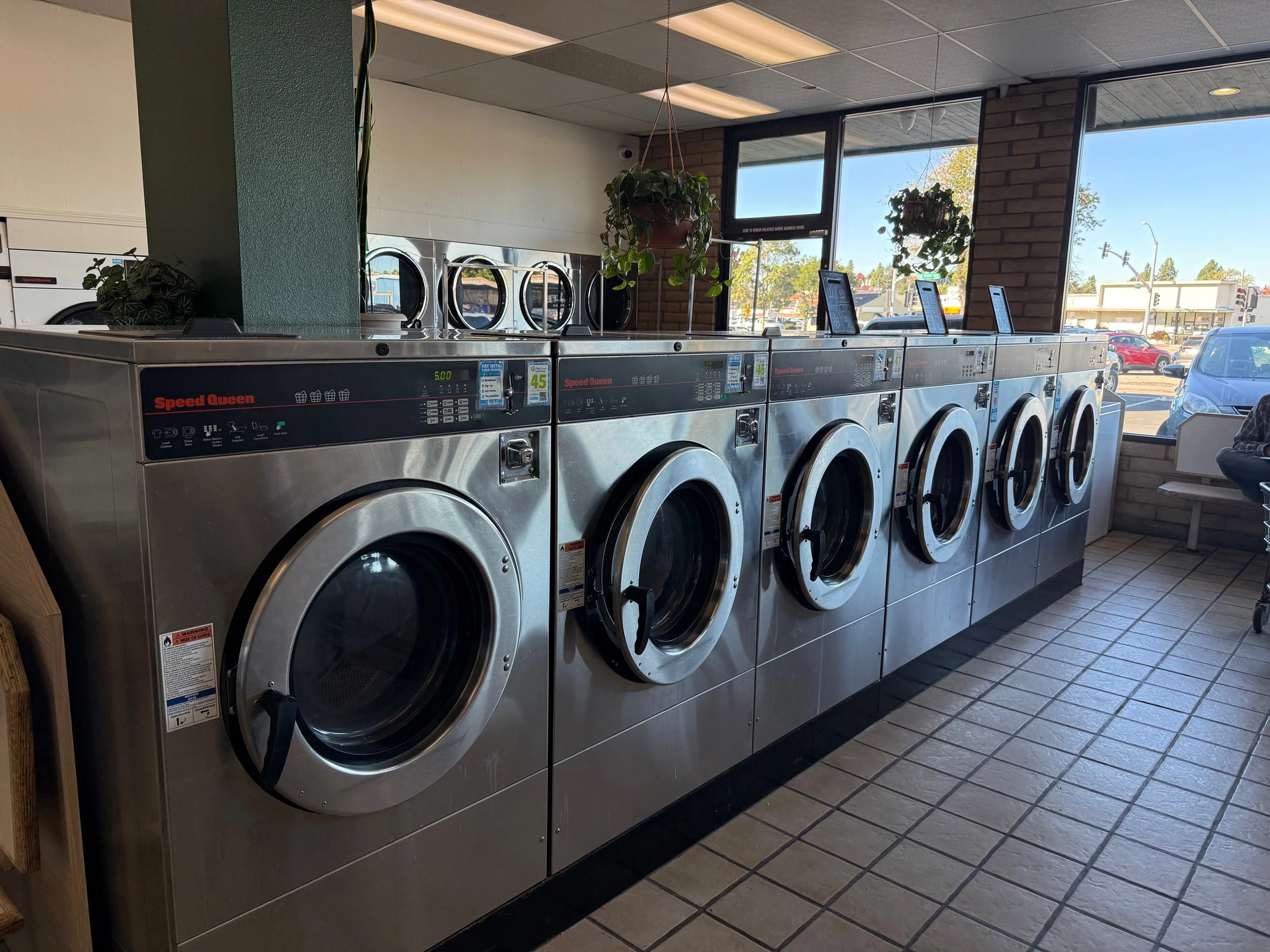 A row of industrial-sized stainless steel washing machines in a laundromat with a tiled floor, a brick wall, large windows, and some potted plants on top of the washers.