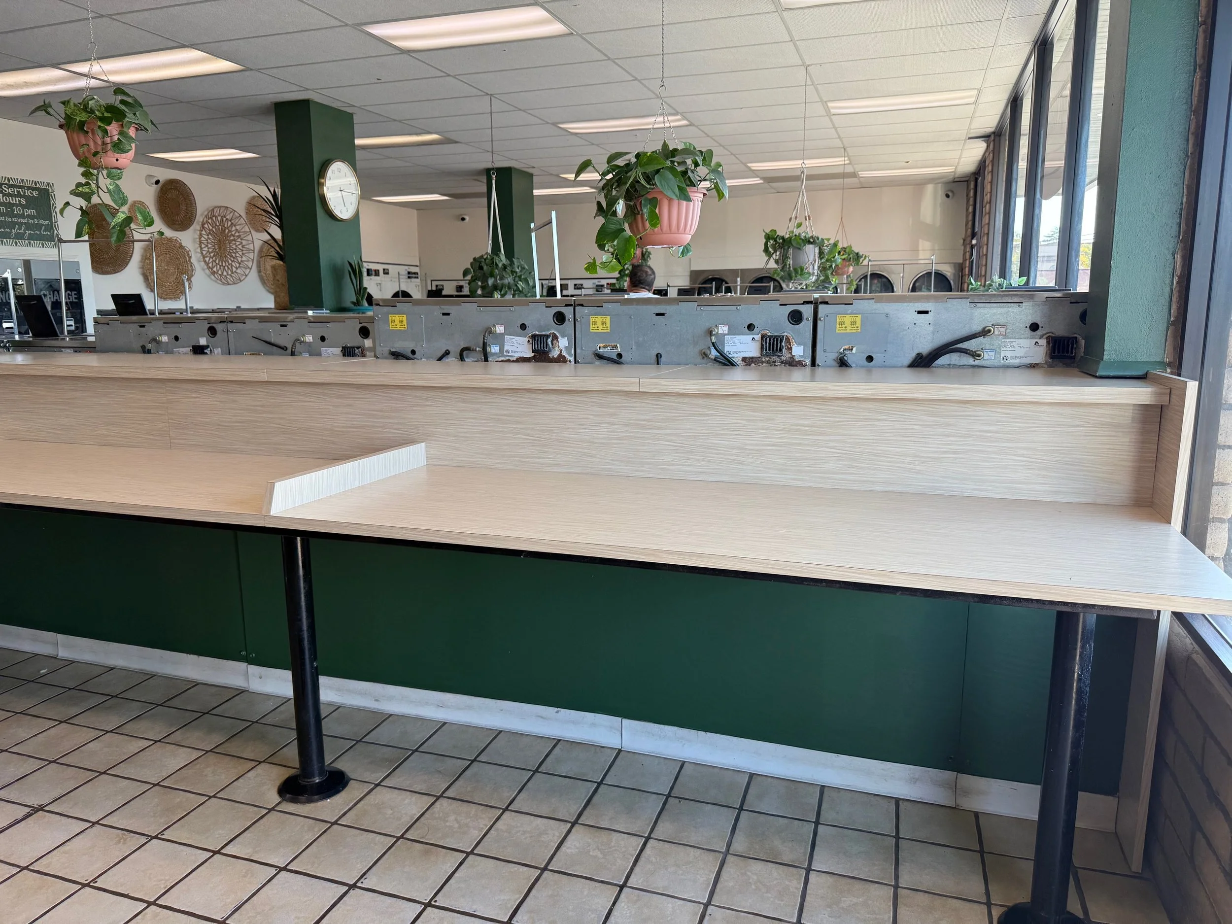 Empty wooden counter with some missing equipment, in a coffee shop or cafe, with green walls and hanging potted plants, and washing machines in the background.