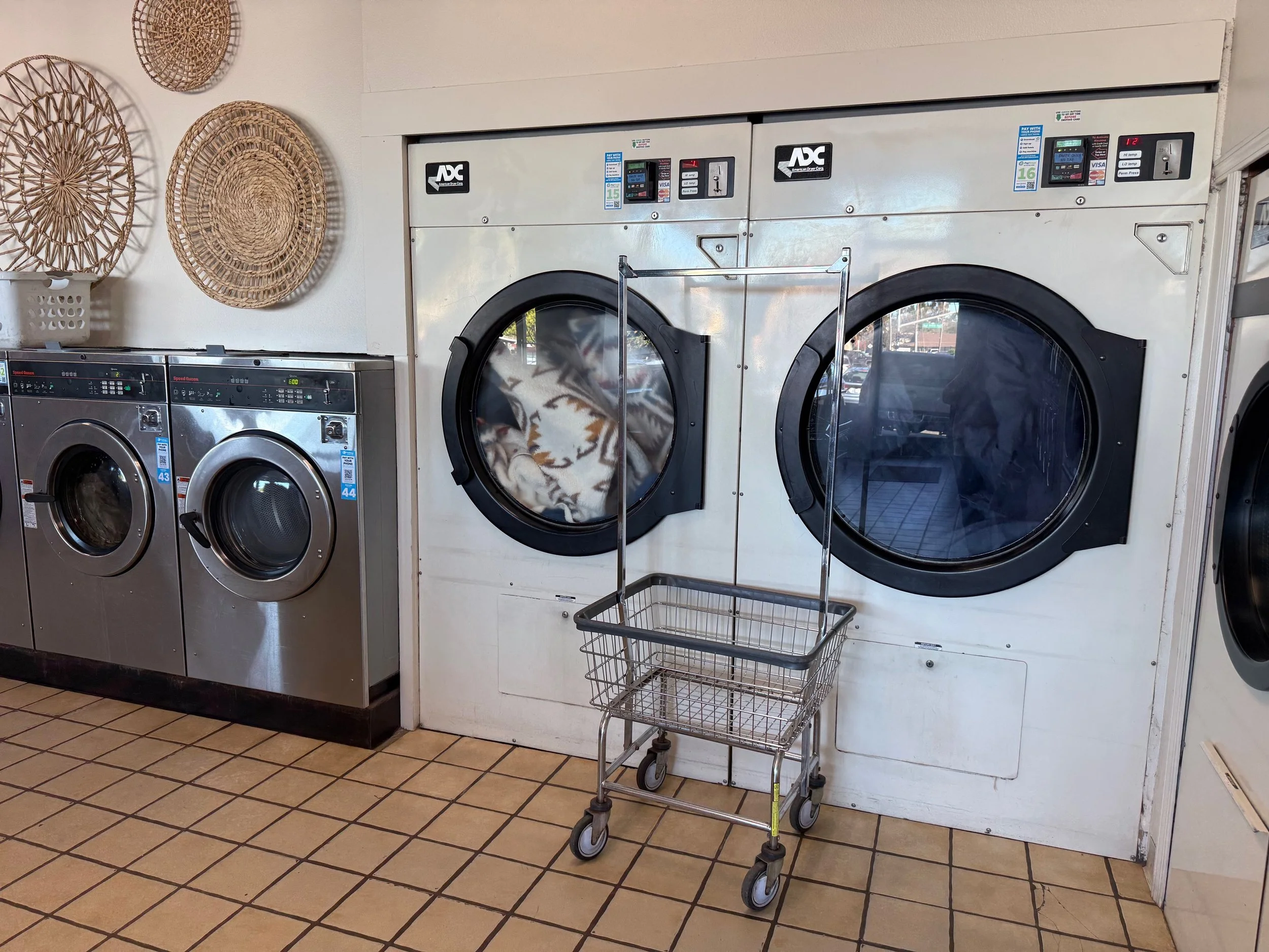 Commercial laundromat with front-loading washing machines, one of which is actively washing clothes. A small laundry cart with wheels is positioned in front of the machines. The flooring consists of tan tiles, and basketly woven wall decorations are visible on the wall to the left.