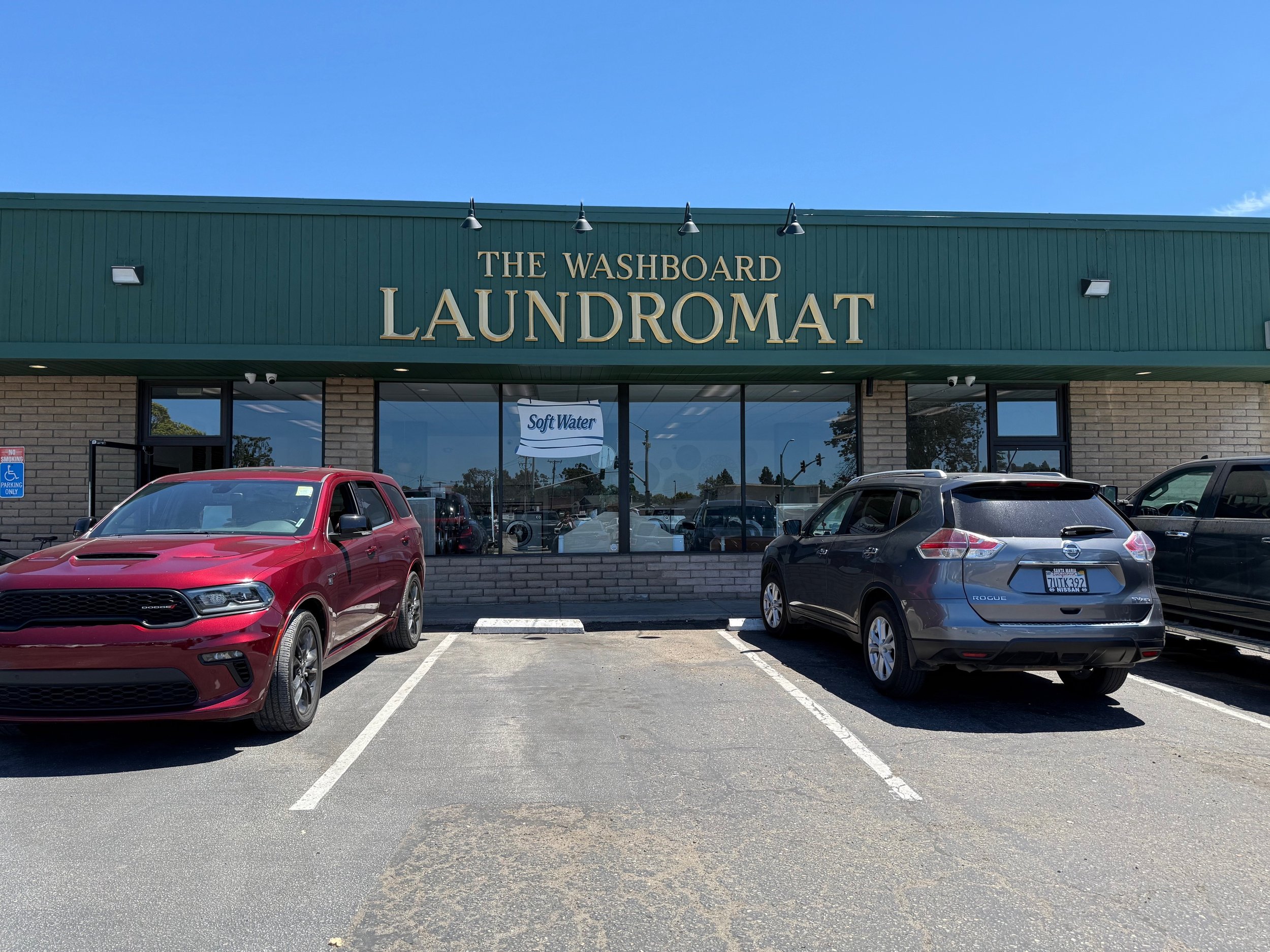 The exterior of a laundromat called 'The Washboard Laundromat' with several parked cars in front, including a red SUV and a blue hatchback, under a blue sky.