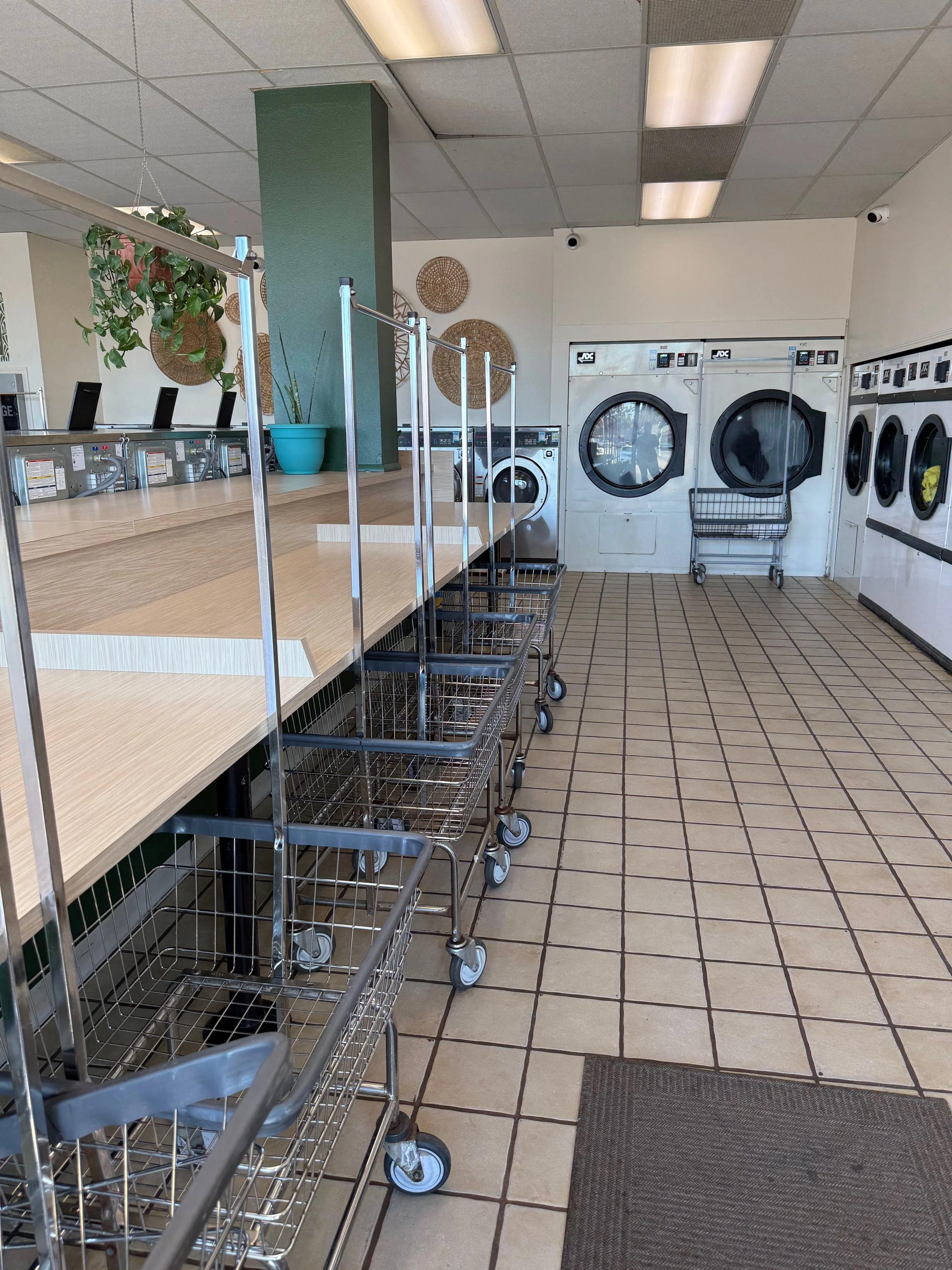 Empty laundromat with washing machines and dryers, shopping carts along a counter, and decorative woven wall art.
