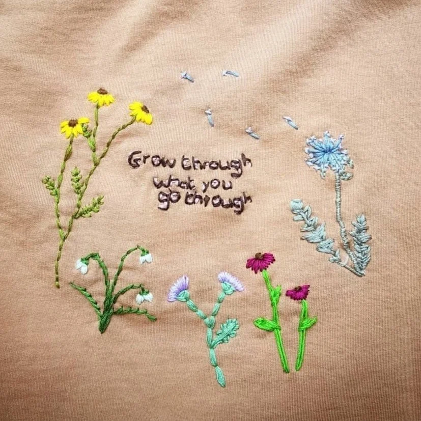 Embroidery of various colorful flowers on a beige fabric background with the quote "Grow through what you go through" stitched in the center.