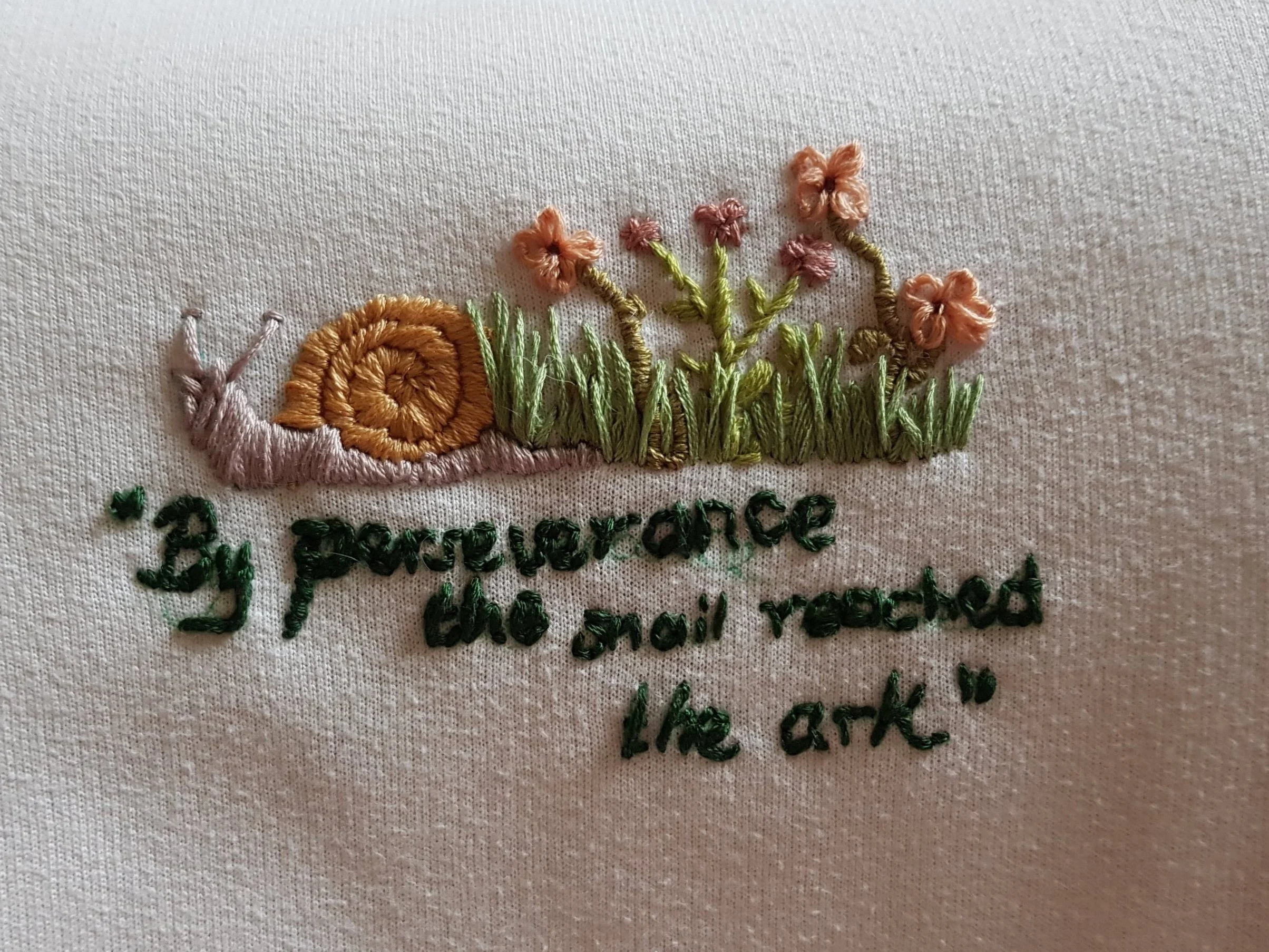 Embroidery of a snail with a shell, grass, and pink flowers, along with the phrase 'By perseverance she kept reached like ark' embroidered underneath.
