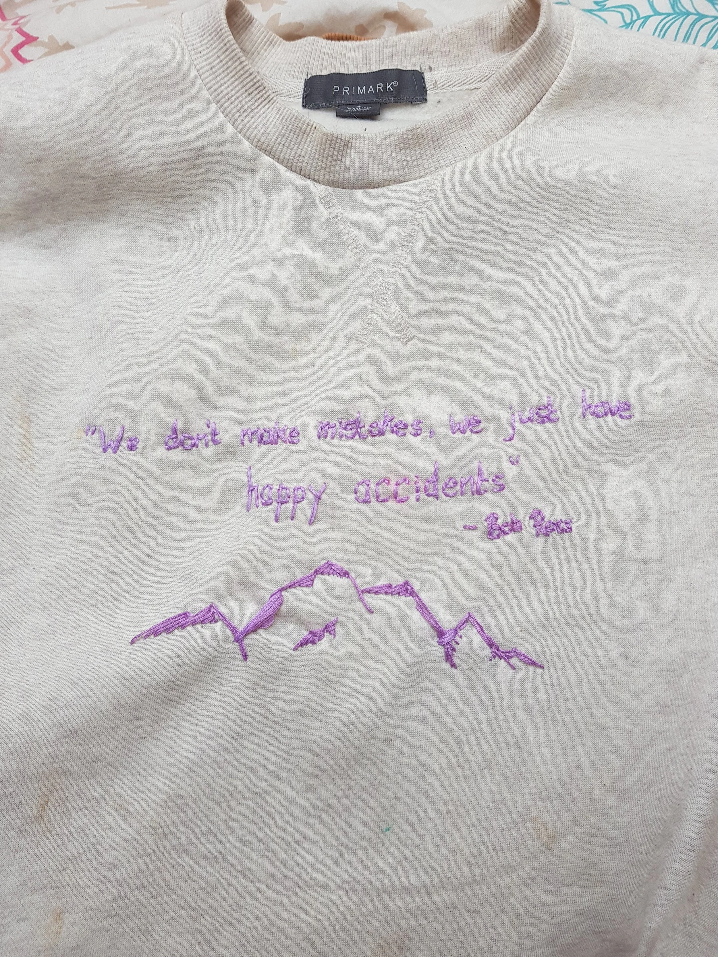 A beige sweatshirt with purple embroidered text that says, "We don't make mistakes, we just have happy accidents" - Bob Ross, and an embroidered mountain range below the text.