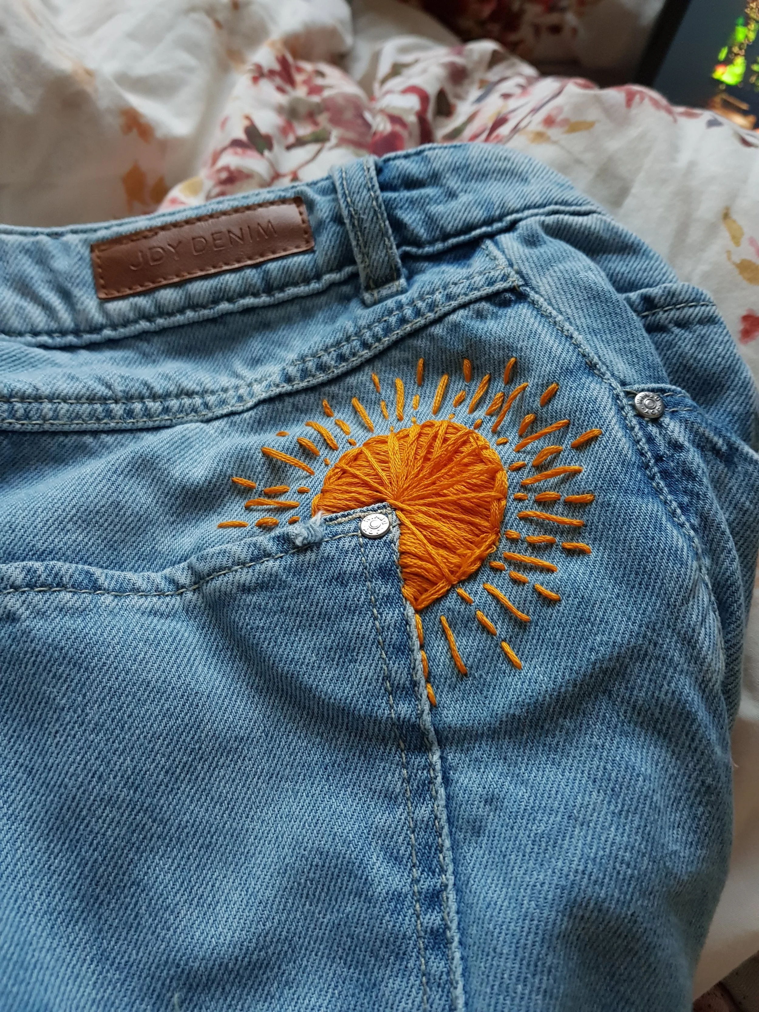 Close-up of light blue denim jeans with a yellow embroidered sun rays design on the back pocket.