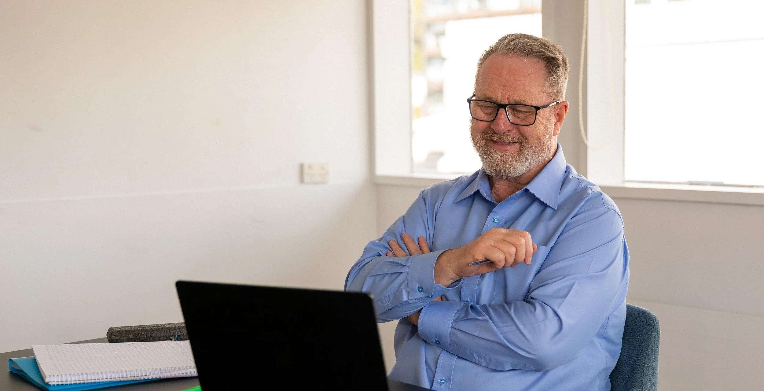 A smiling older man with glasses and a beard wearing a light blue dress shirt, sitting at a desk with a laptop, notebook, and pen, in a bright office with windows.