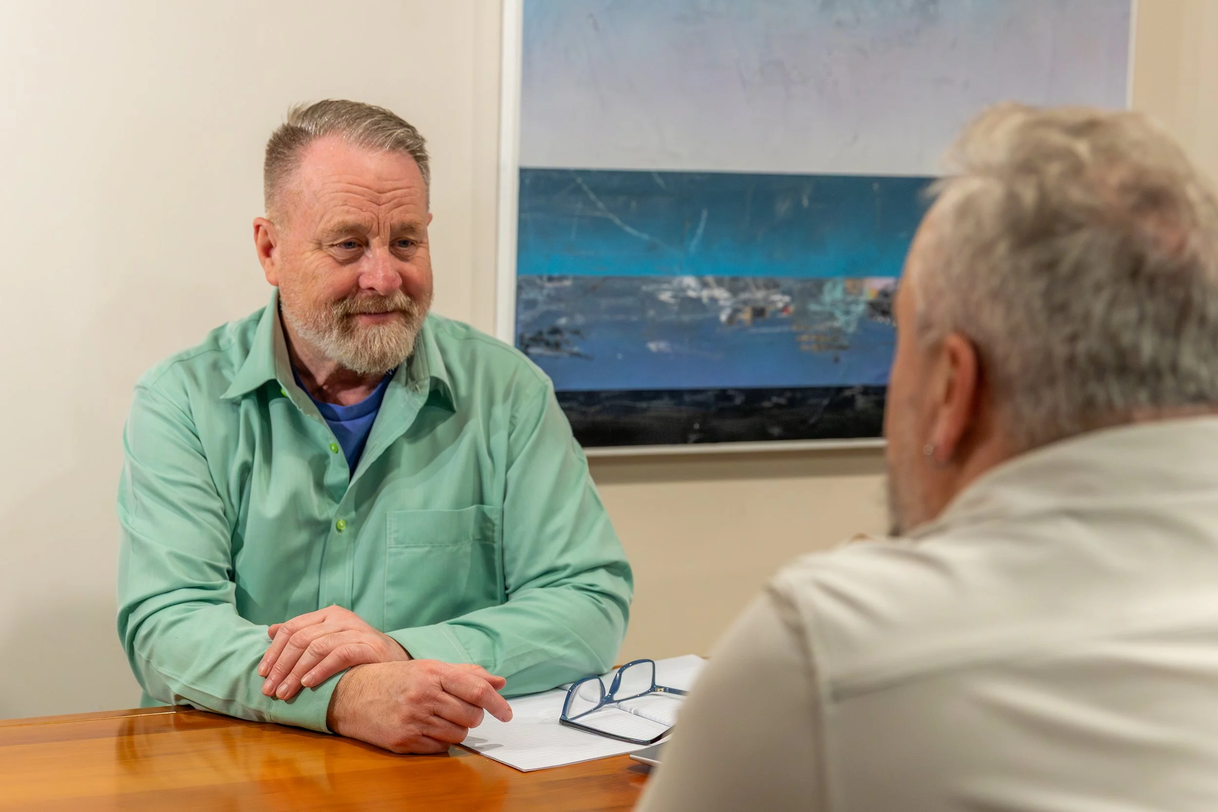 A man with gray hair and a beard wearing a green shirt is sitting at a table, engaging in conversation with an older woman with gray hair seen from behind. There is a pair of glasses and a notebook on the table, and a painting of a seascape hangs on the wall behind.