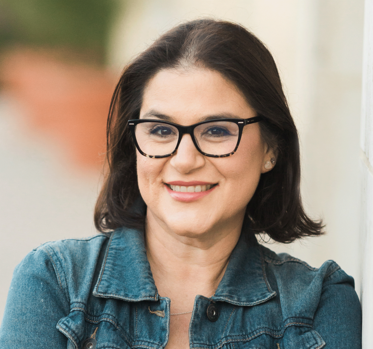 A woman with dark brown hair, wearing glasses and a denim jacket, smiling outdoors.