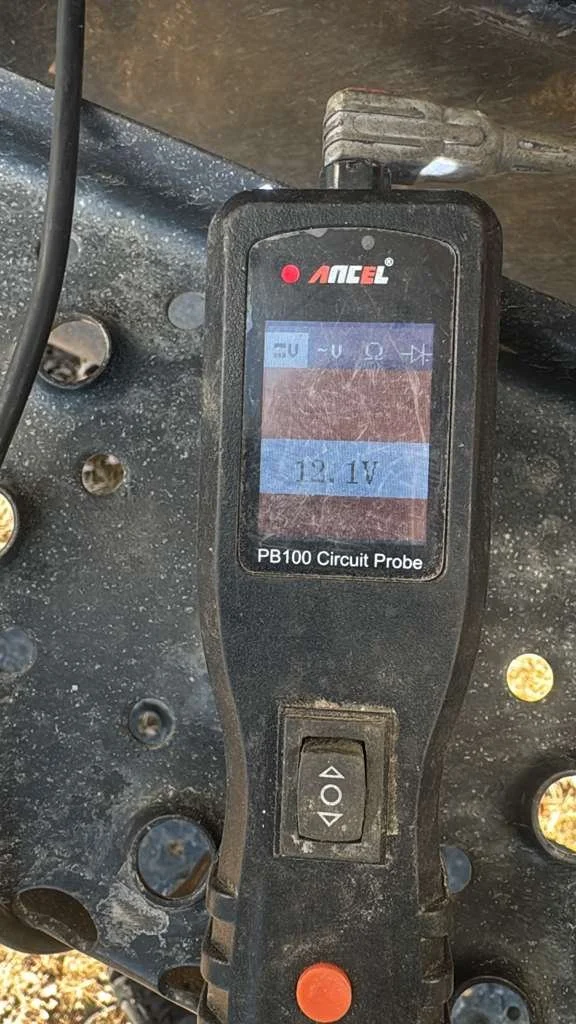 A circuit probe device showing a voltage of 12.1V, placed on a dusty, black surface with several holes and some screws.