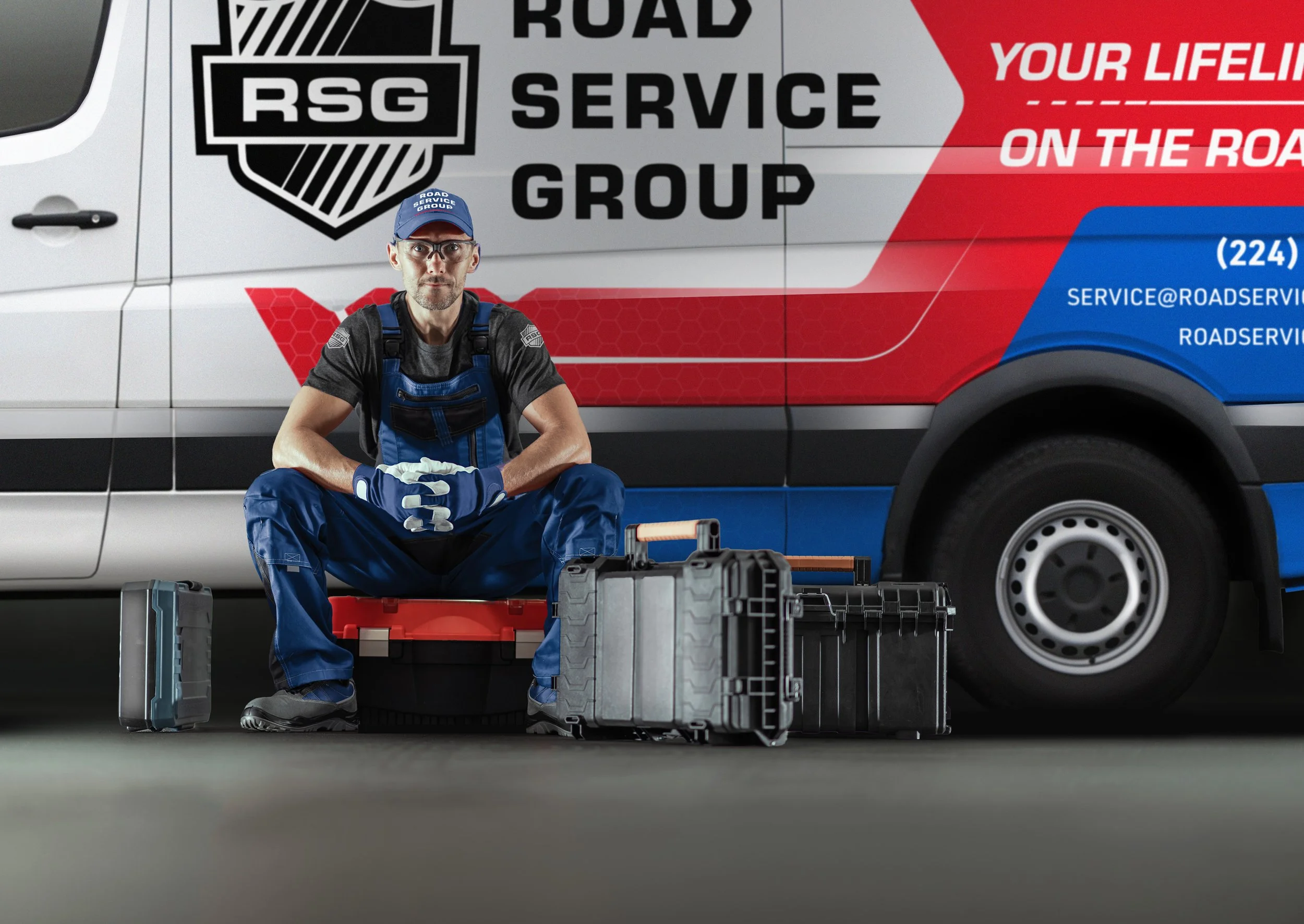 A male roadside assistance worker dressed in blue overalls and gloves, sitting on a black tool case with other tool boxes nearby, in front of a roadside service vehicle labeled 'Road Service Group' with professional contact information displayed on it.