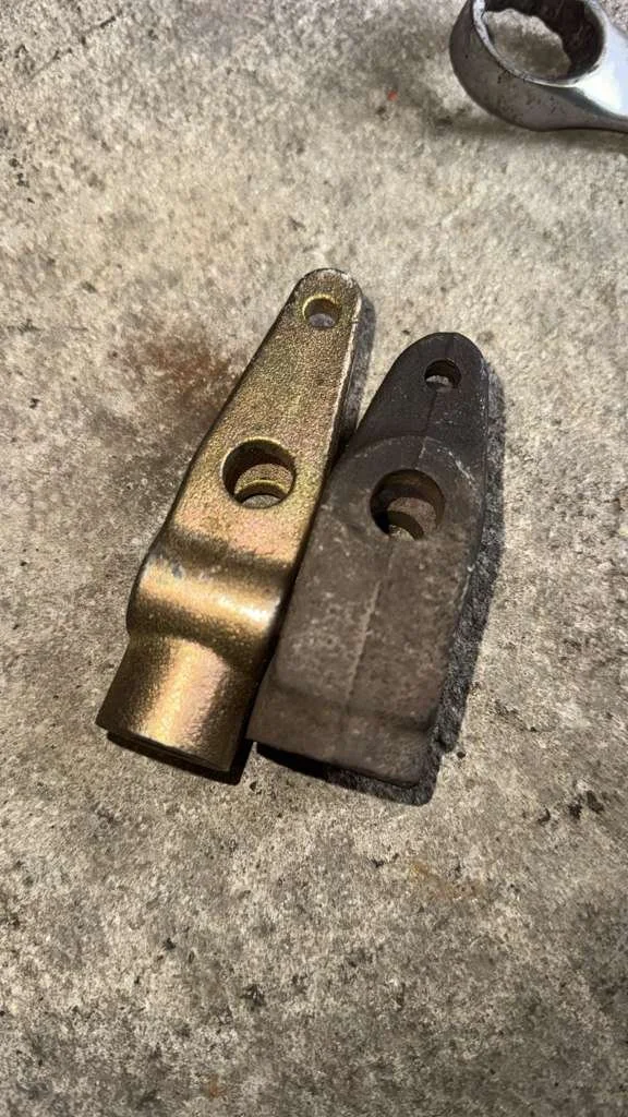 Two metal hinge plates, one new and shiny gold-colored, the other old and worn with rust and corrosion, lying on a concrete surface.