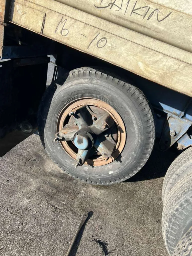 A close-up of a worn-out vehicle tire and rusty wheel rim on a black vehicle, parked on asphalt with a wooden board visible above, bearing handwritten notes and a date.