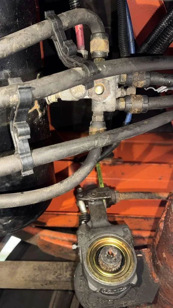 Close-up view of a mechanical system with intersecting metal pipes, fittings, and a hydraulic component, all covered in dirt and grime, with an orange background.